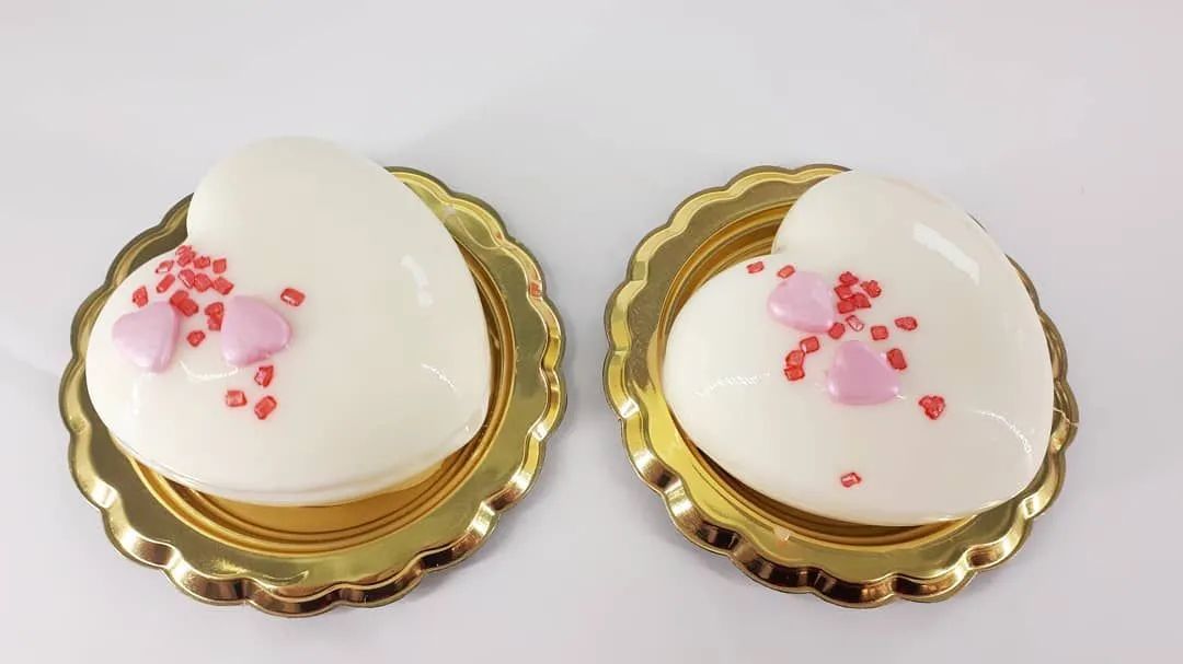 Due pasticcini a forma di cuore, ricoperti di glassa bianca e decorati con piccoli cuoricini rosa e zuccherini rossi, poggianti su basi dorate smerlate.