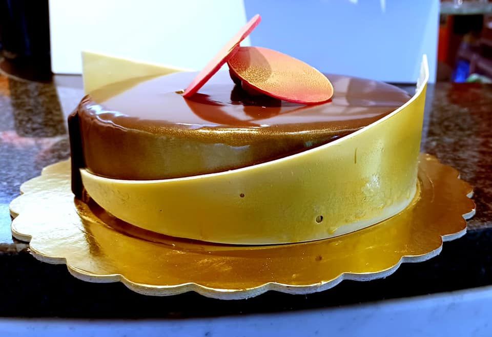 Una torta lucida di mousse al cioccolato su una base dorata smerlata, decorata con scaglie curve di cioccolato bianco e un disco rosa.