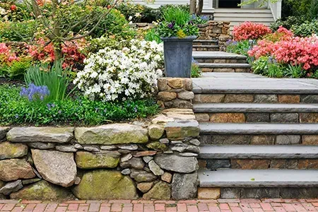 A stone wall with stairs leading up to a garden with flowers.