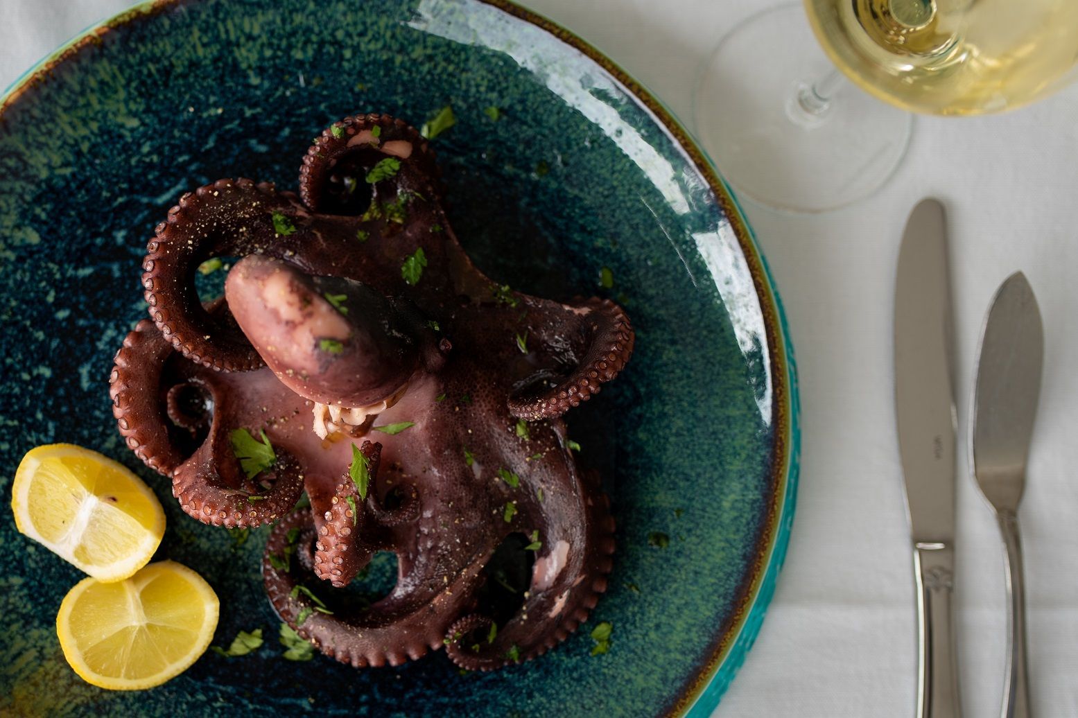 Plated octopus dish on a blue plate with lemon wedges and cutlery on a white table