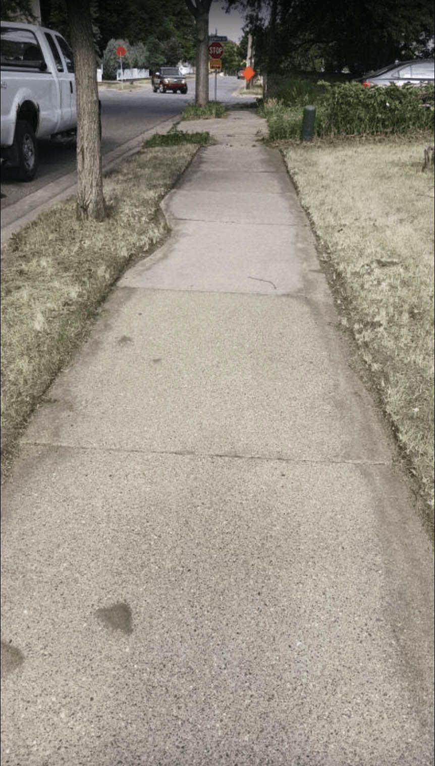 Sidewalk with a tree on one side, a grassy area, and a street with cars in the background.