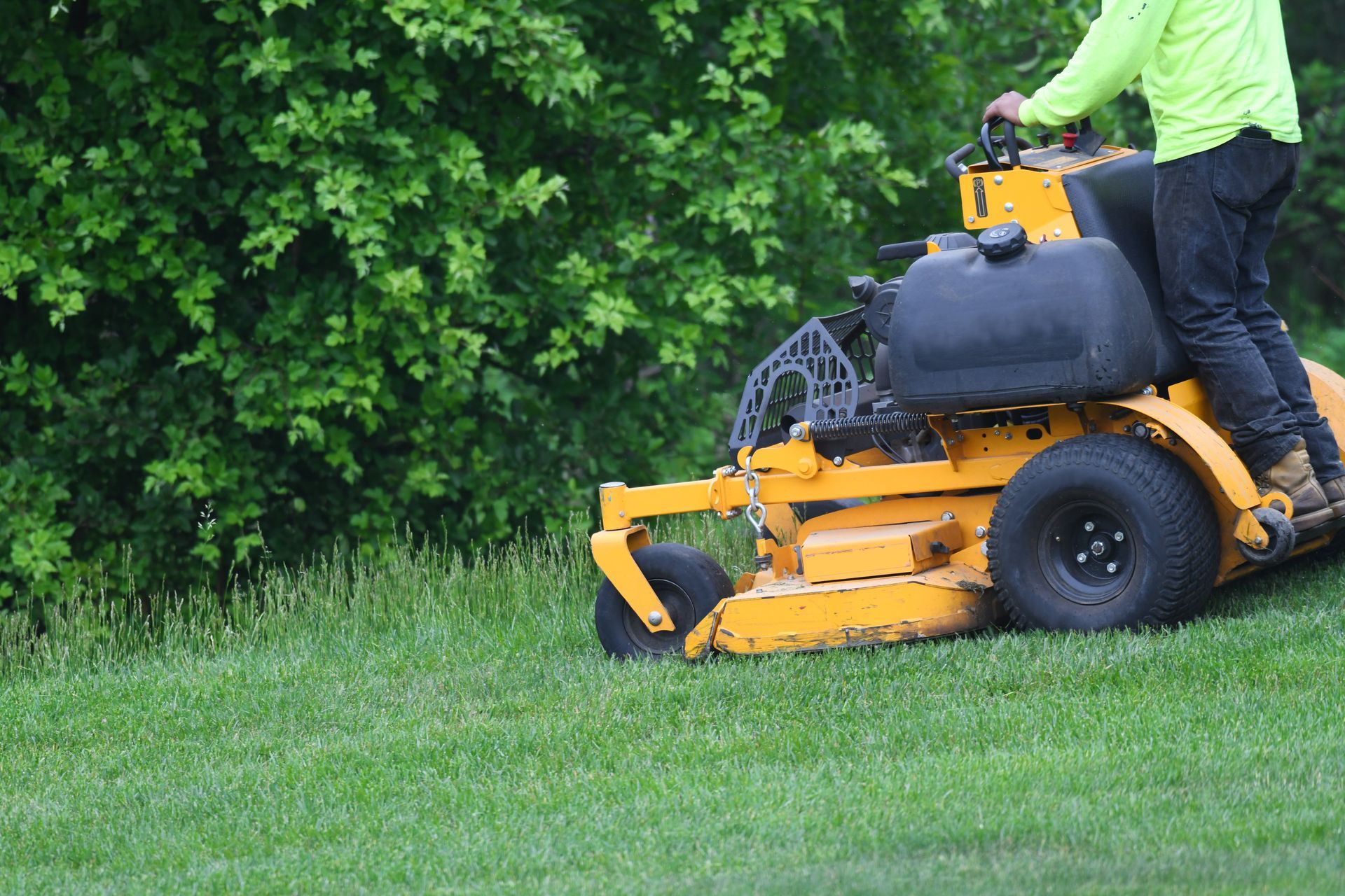 Yellow riding lawnmower on a green lawn, next to a green bush. A person in yellow shirt is operating the mower.