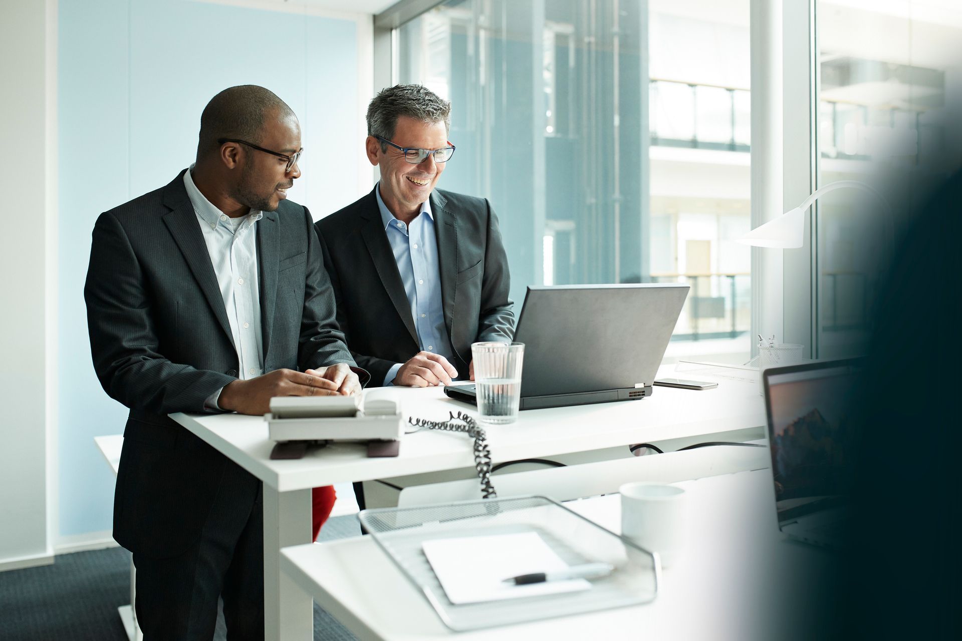 Two men are sitting at a desk looking at a laptop computer.