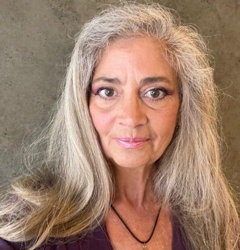 A woman with long gray hair is wearing a purple shirt and a black necklace.