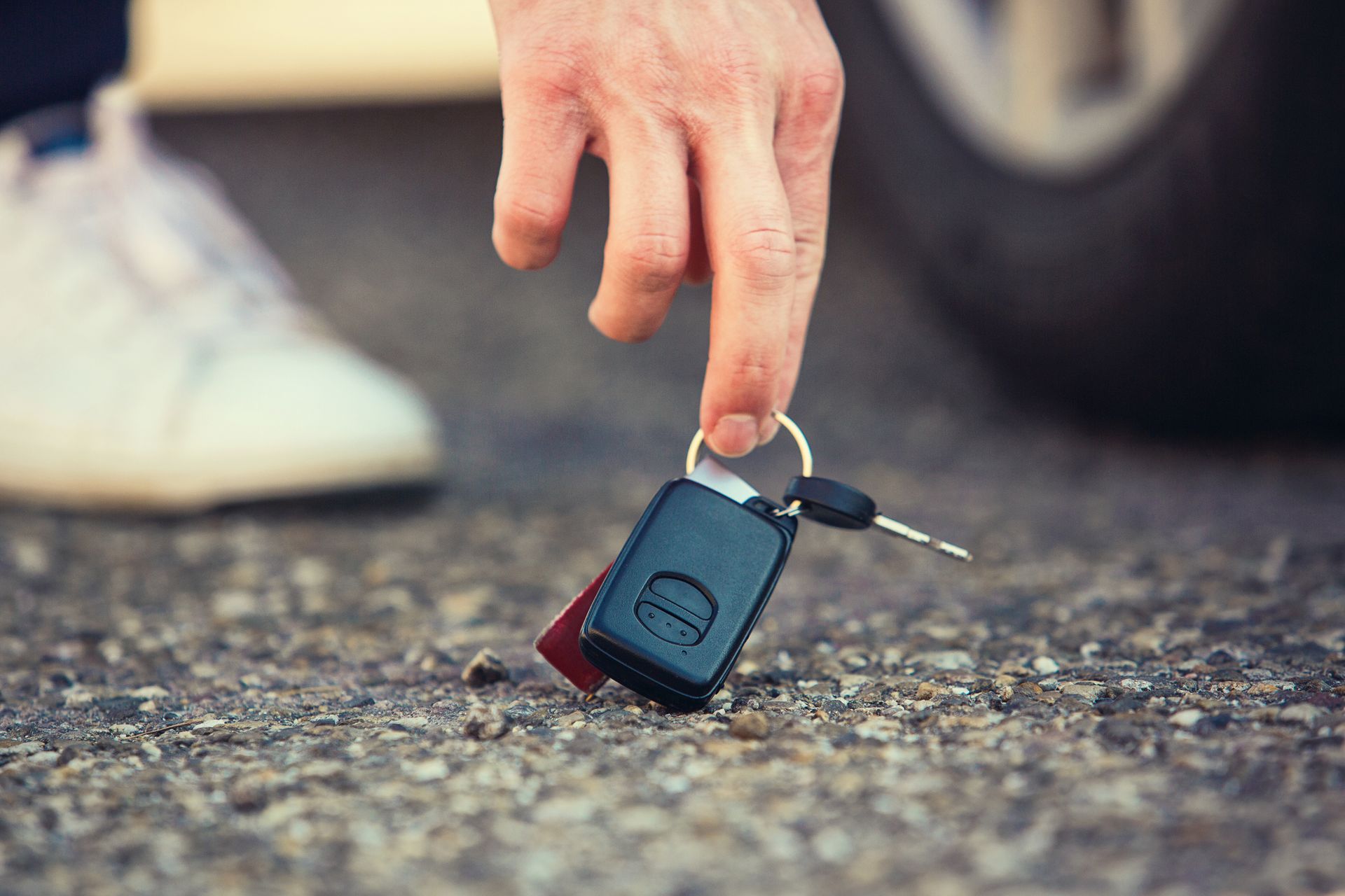 Hand reaching for car keys lying on the ground near a tire and white shoe.