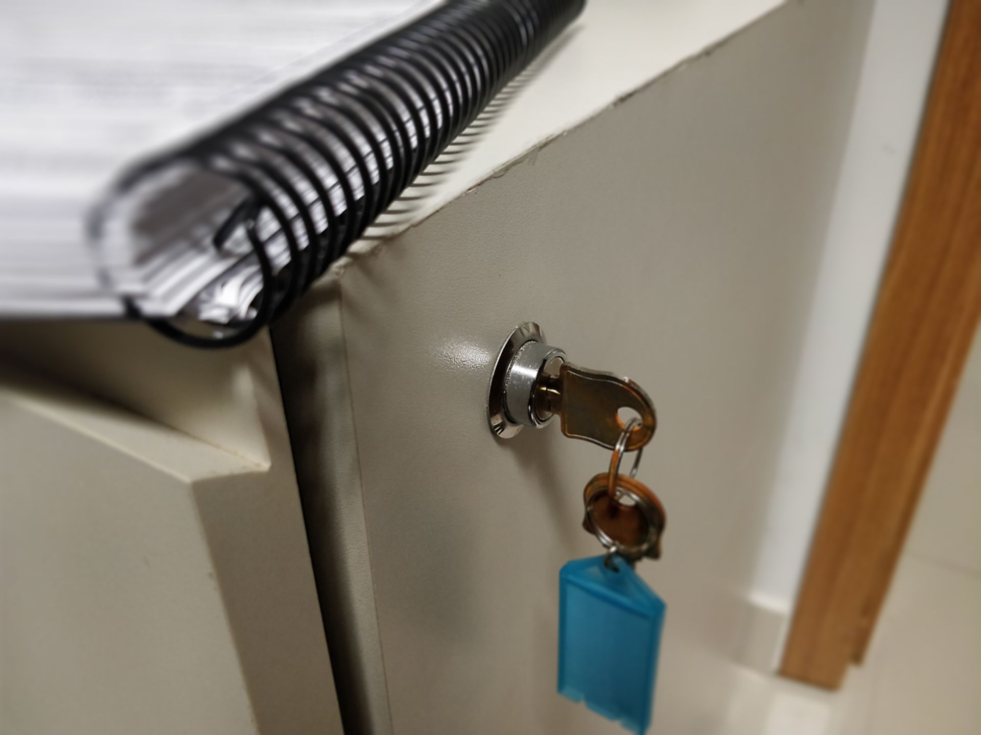 Key in a locked cabinet, with blue tag. A spiral-bound notebook rests on top.