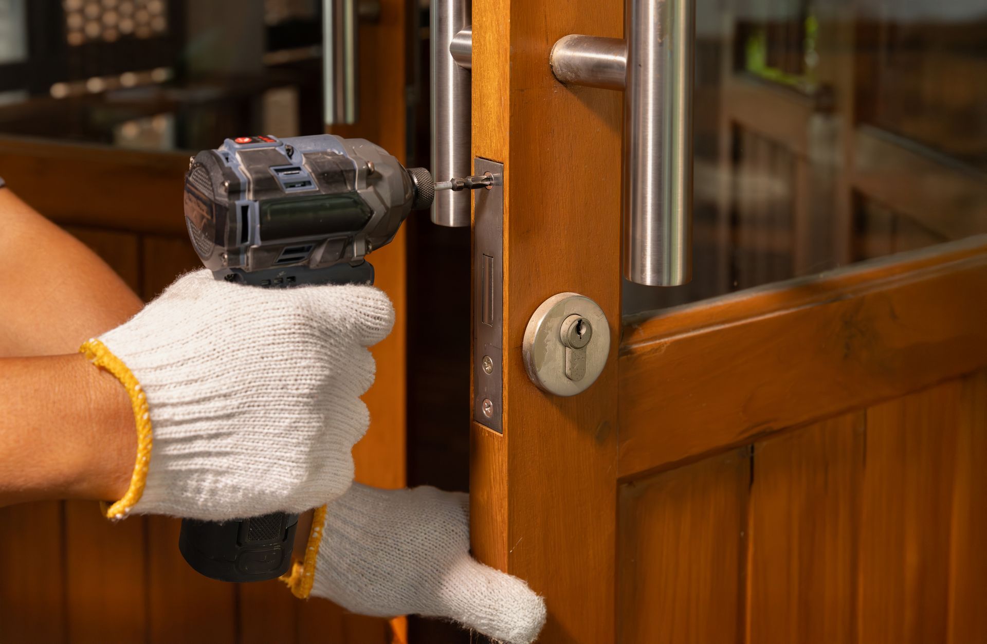 Person in gloves using a power drill to install a door latch on a wooden door.
