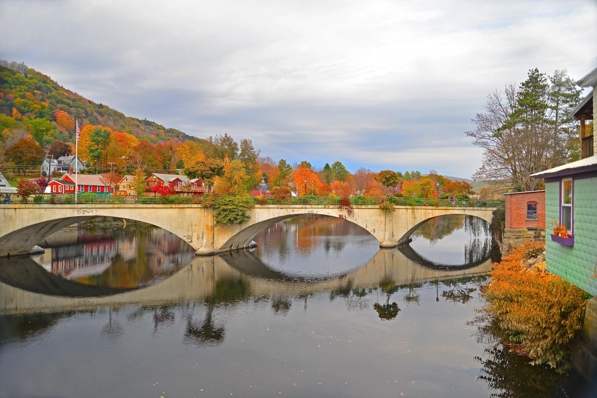 Stone arch bridge over calm water, fall foliage and buildings in background.