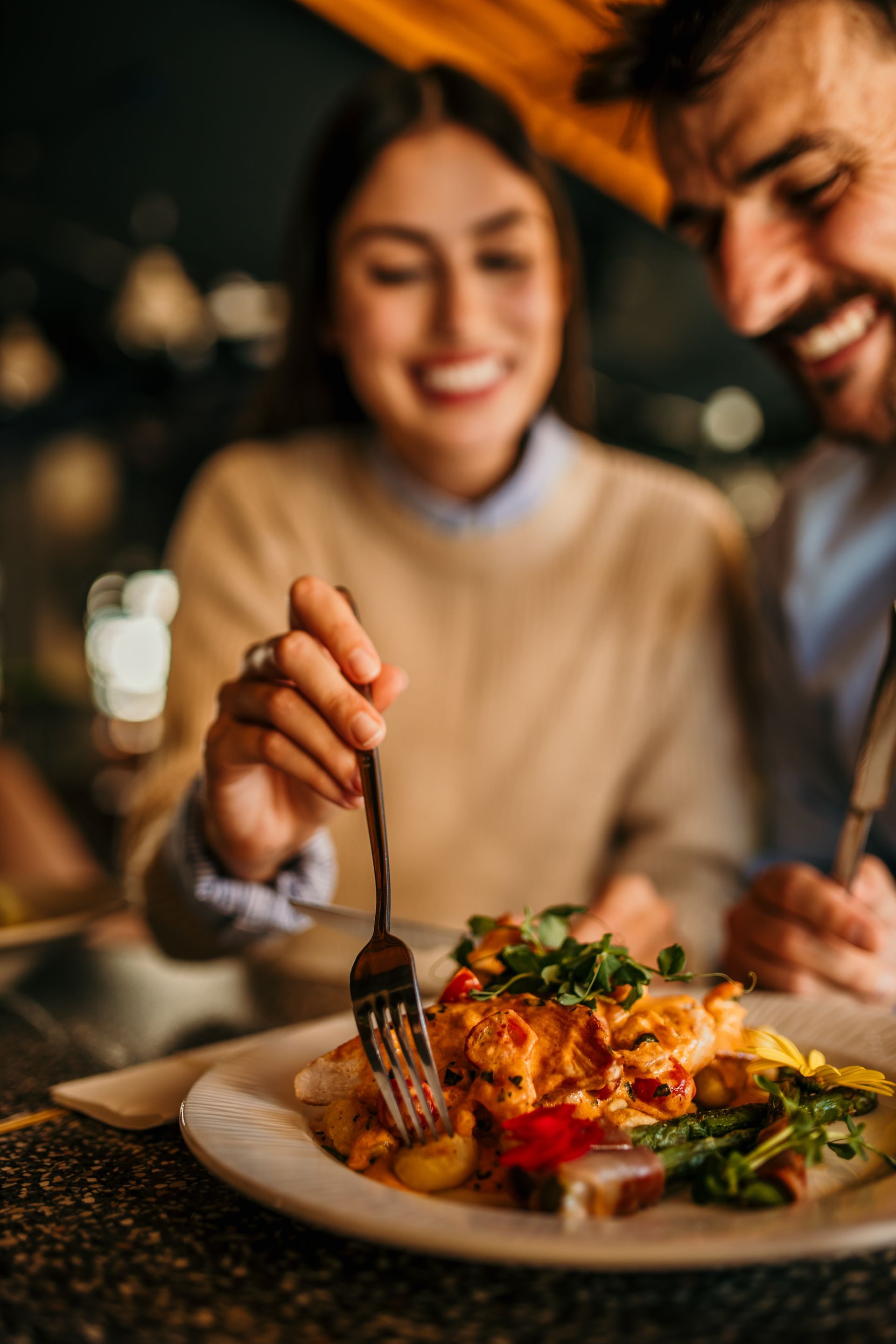 Couple smiling while eating a meal at a restaurant. A woman uses a fork.