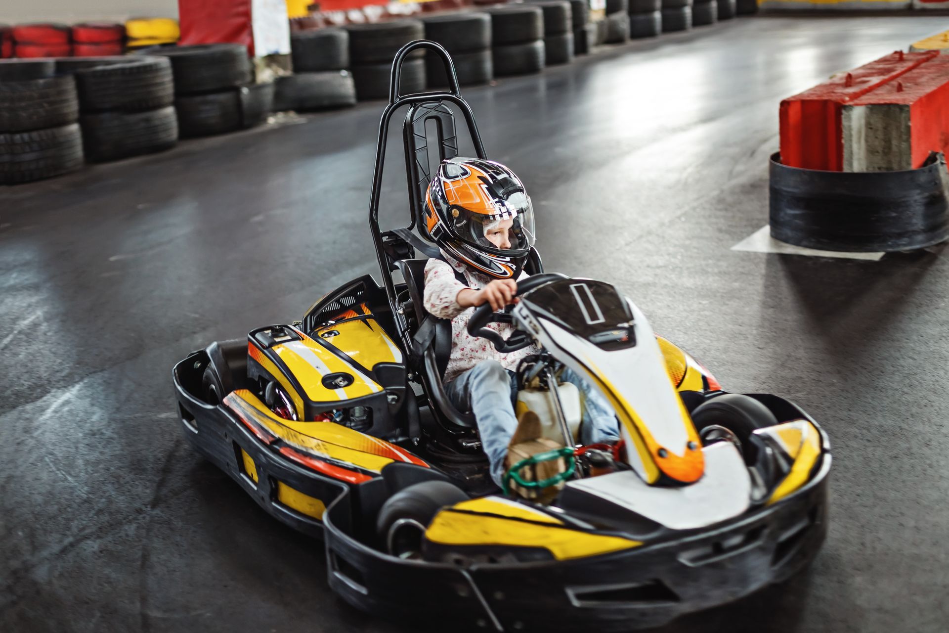 Young child in a yellow and black go-kart, wearing a helmet, on an indoor track.