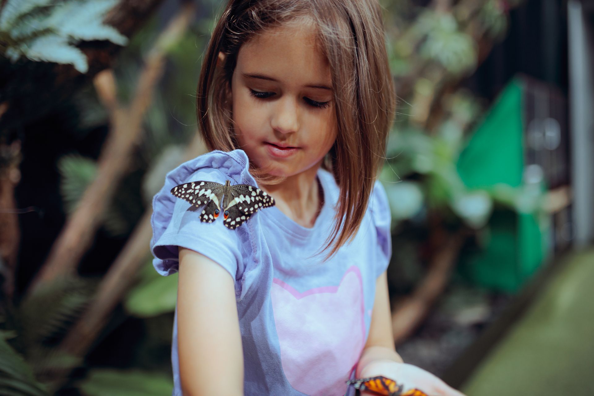 A young person in a blue shirt with a butterfly perched on their shoulder in a lush, green garden setting.