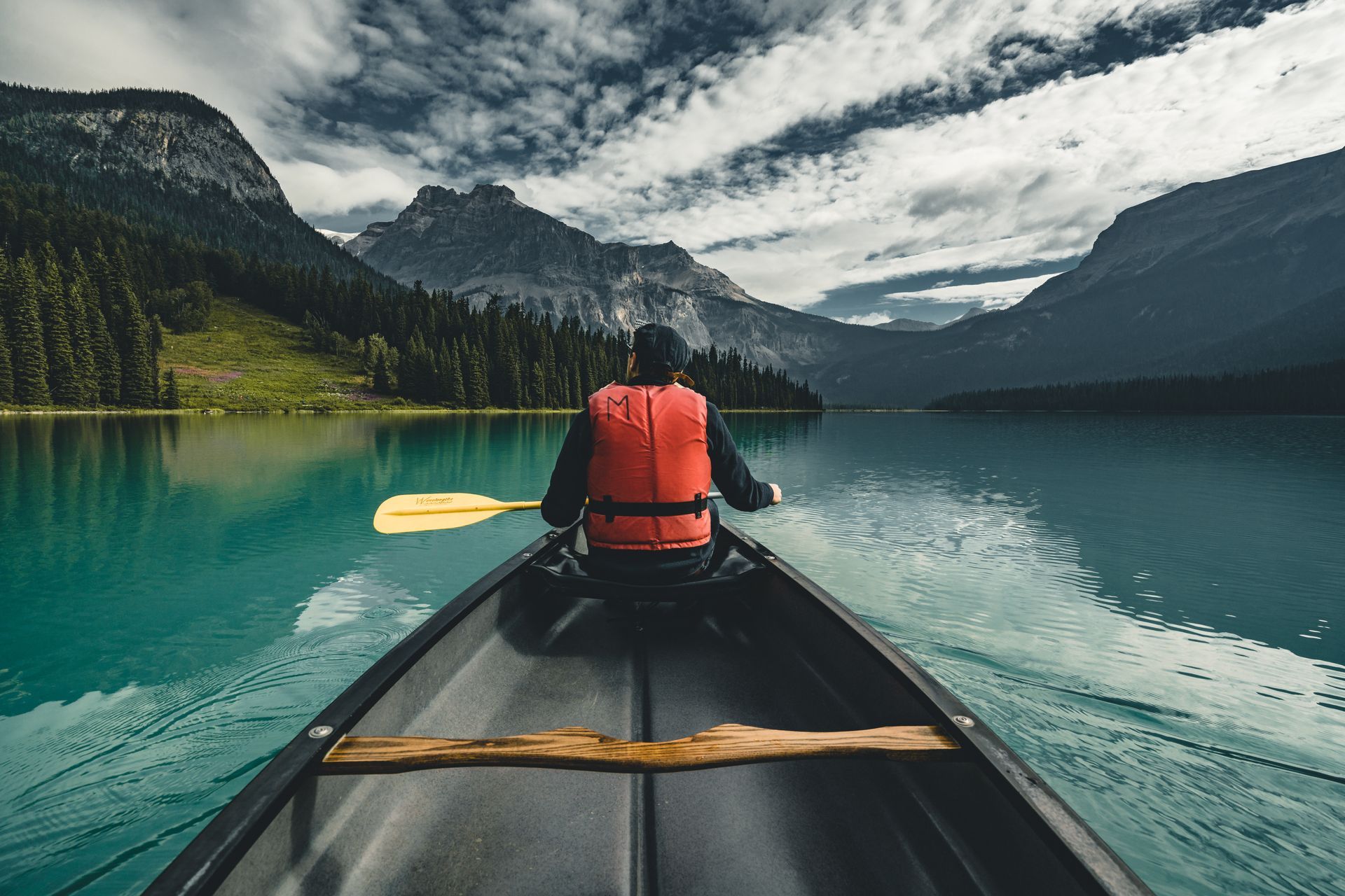 Person in an orange life vest paddles a canoe on a turquoise lake, mountains in the background.