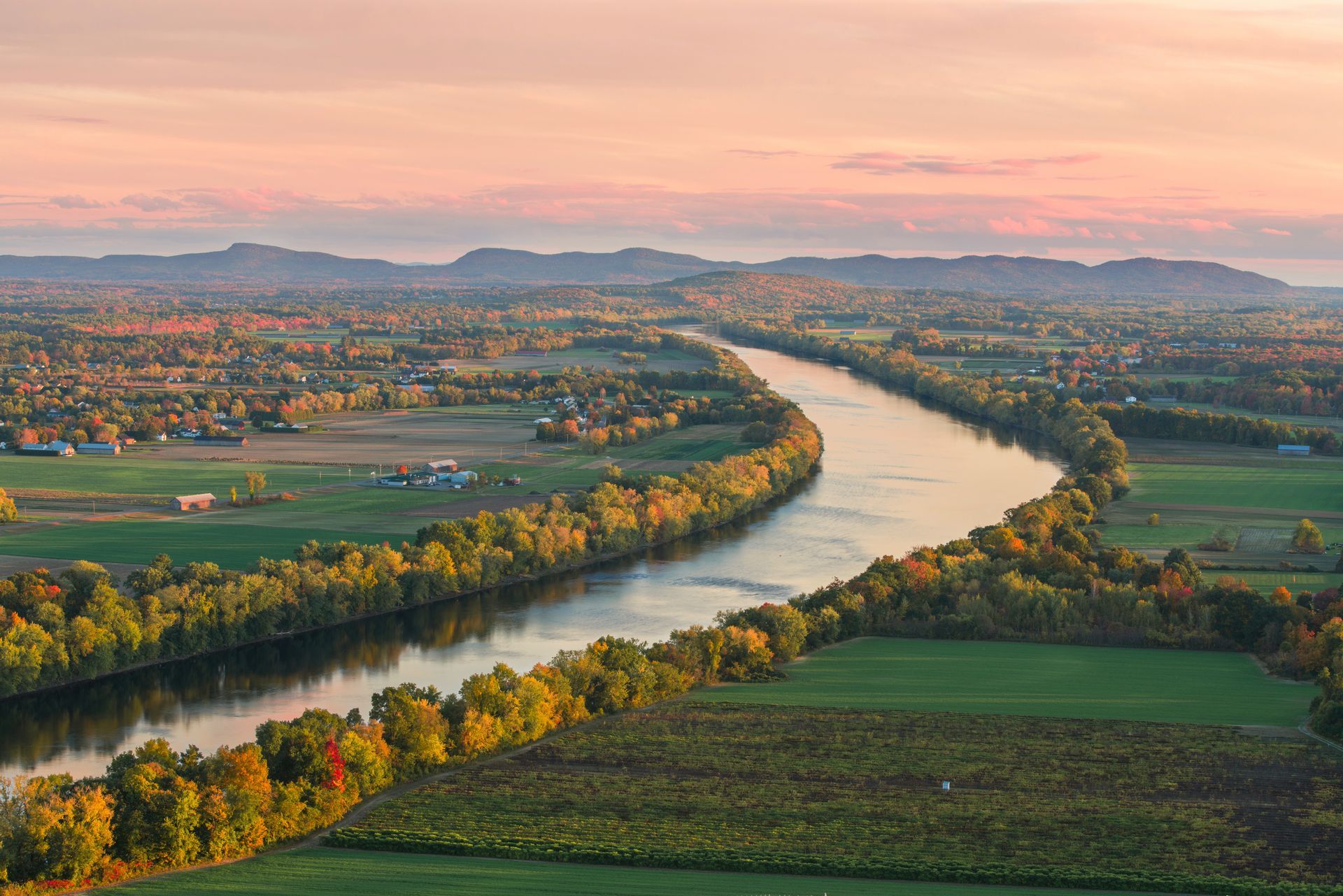 Winding river through green fields at sunset, with autumn trees and distant mesas under a pink sky