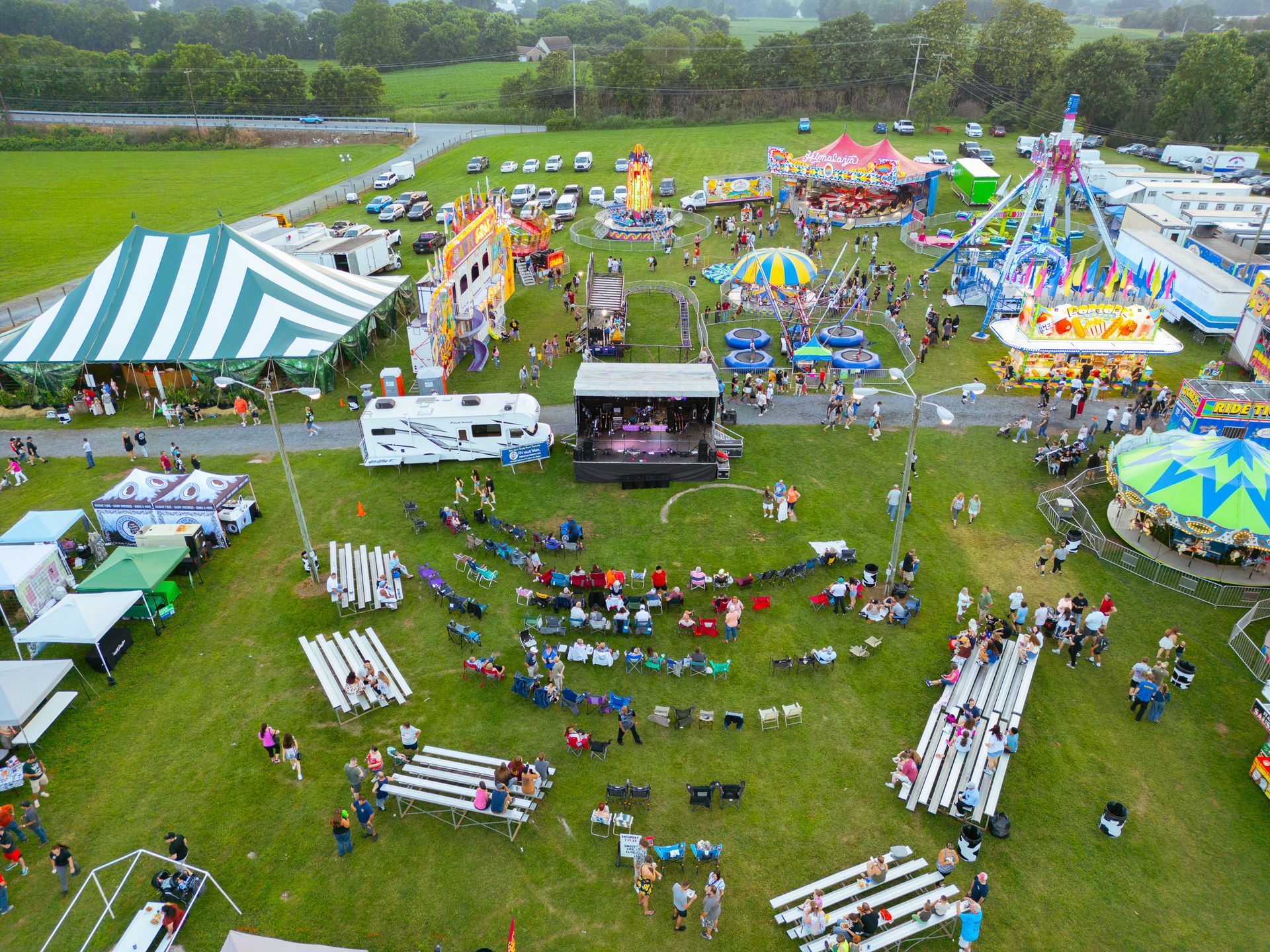 Aerial view of a fair with rides, tents, a stage with a band, and people on a grassy field.