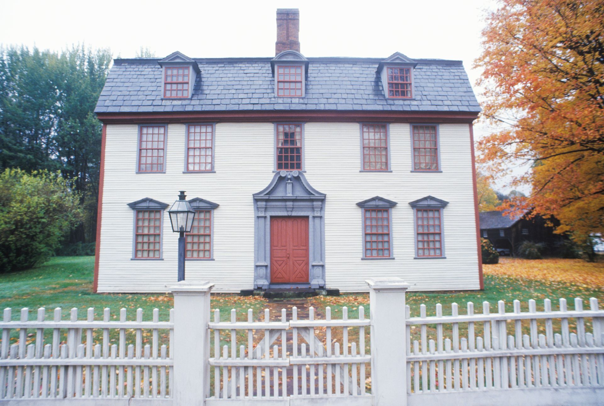 A two-story, white colonial-style house with a red door, a gambrel roof, and a white picket fence in front.