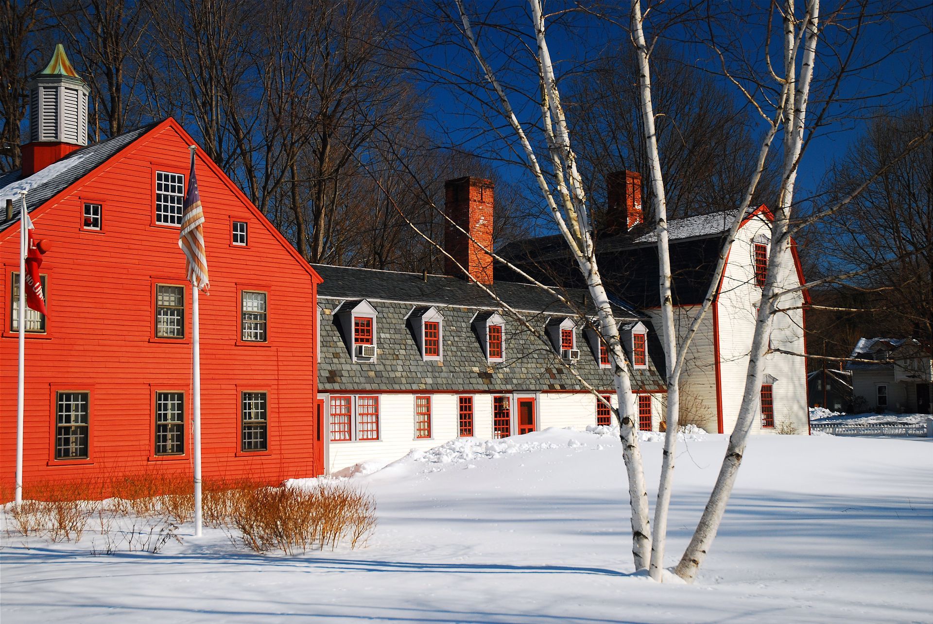A red clapboard building with a white wing stands in deep snow on a sunny winter day with a tall white birch tree nearby.