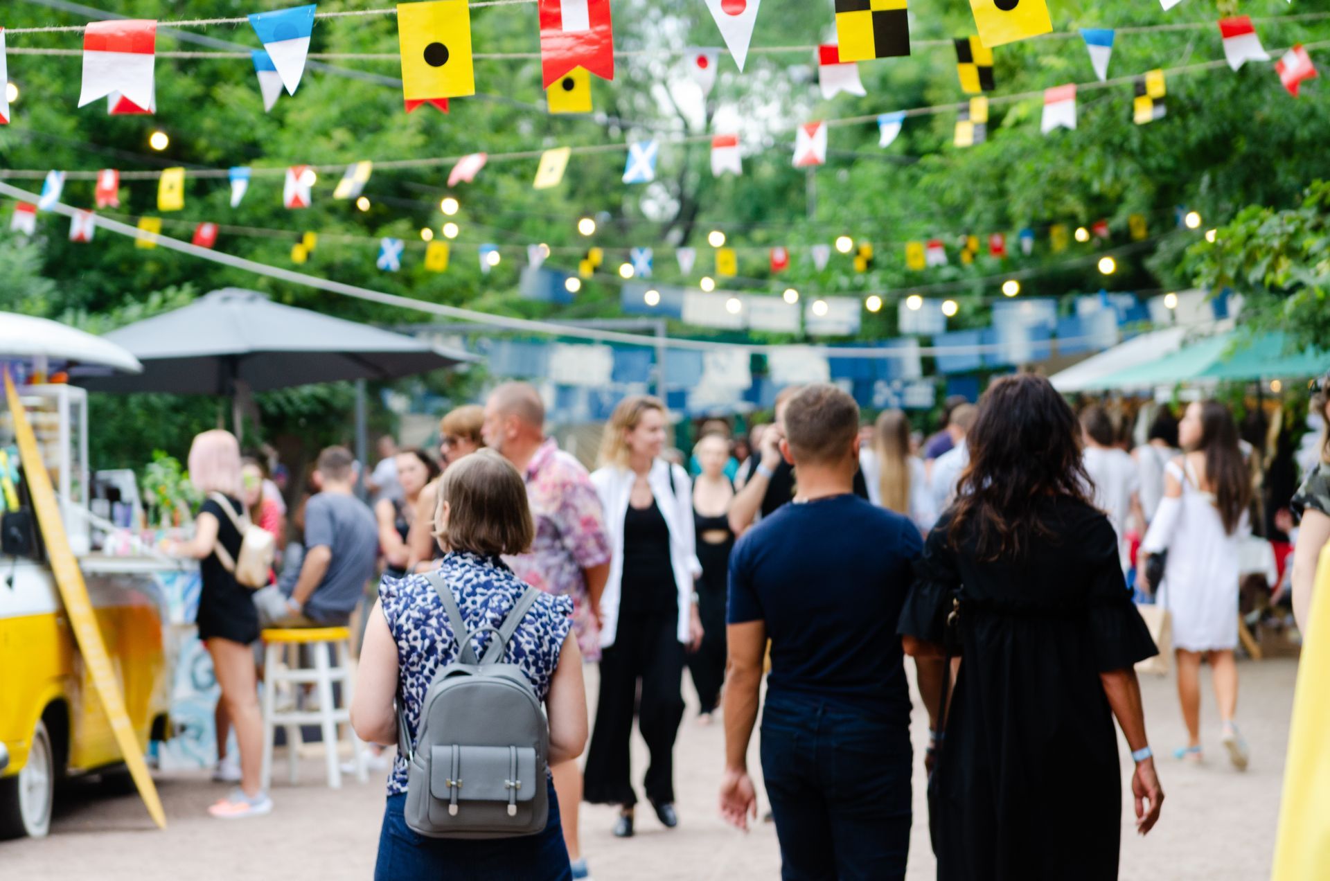 People at an outdoor event, with flags strung overhead. Food stalls and a yellow van are visible.
