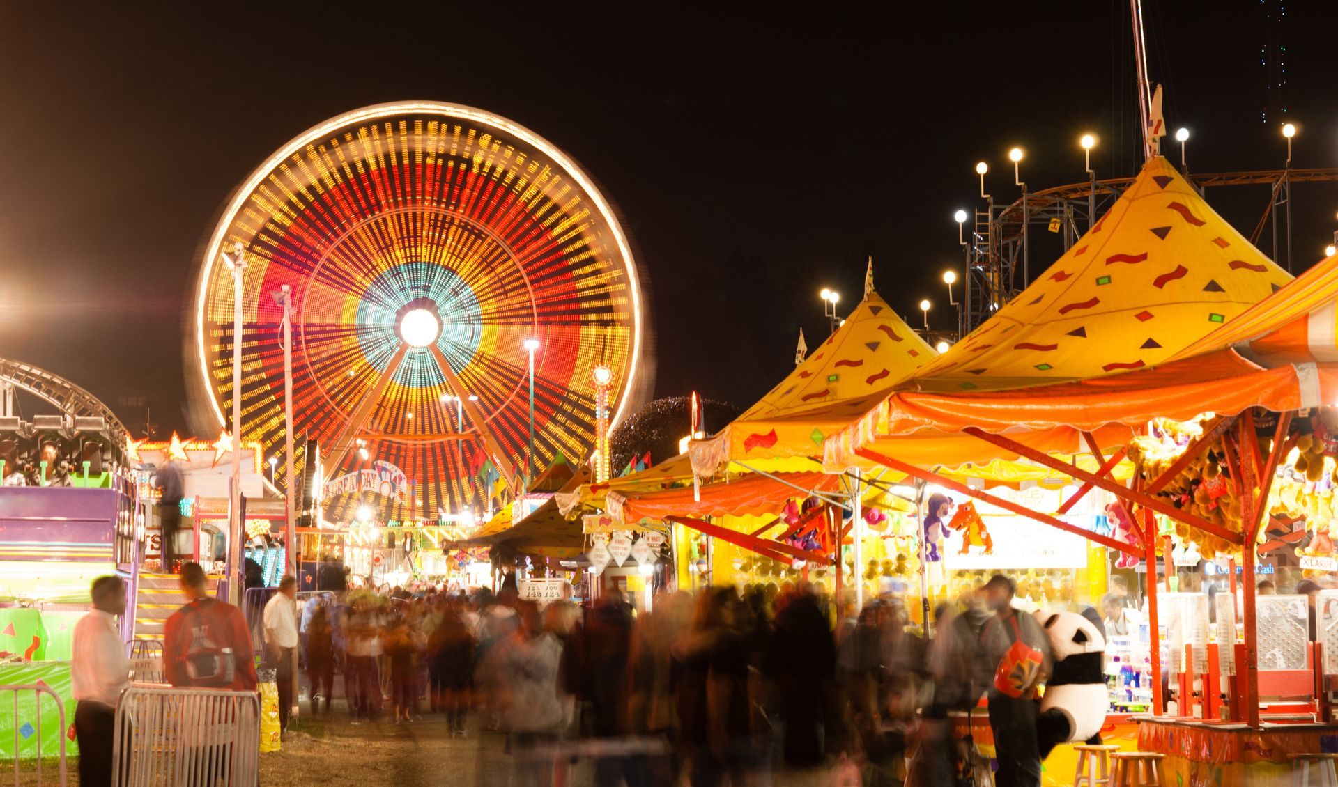 Nighttime carnival scene with a bright, spinning Ferris wheel and crowded food stalls.