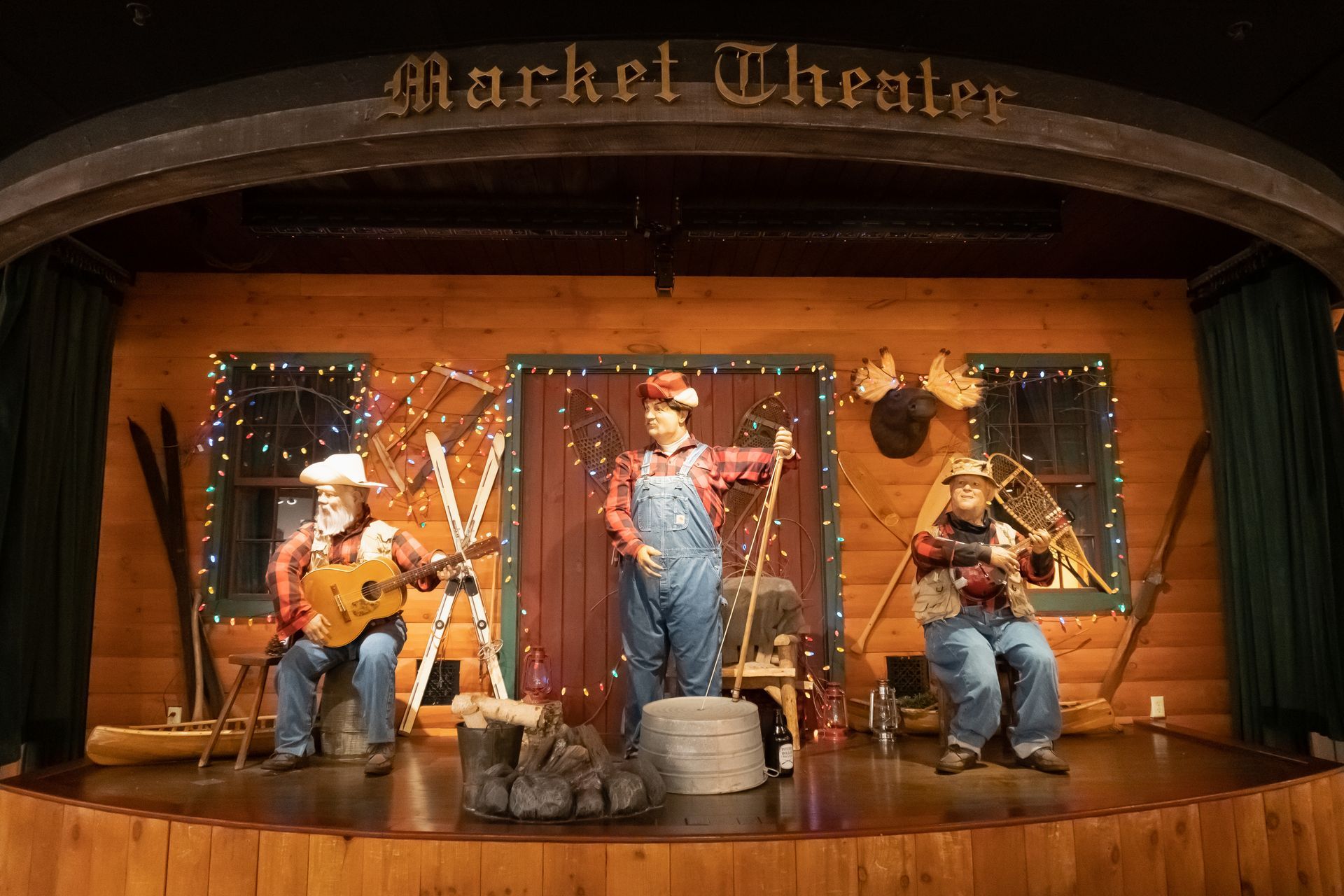 Three performers on a rustic, log-cabin-themed Market Theater stage with props including skis, a moose head, and lights.