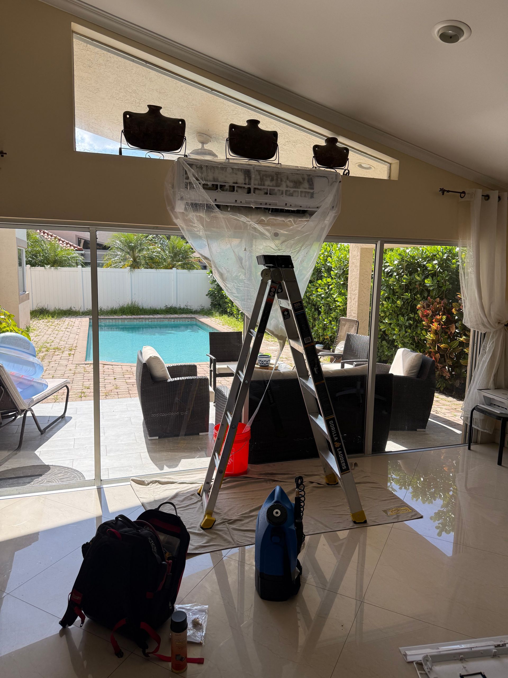 A mini-split AC unit, covered in protective plastic sheeting, is being serviced with a ladder positioned underneath.