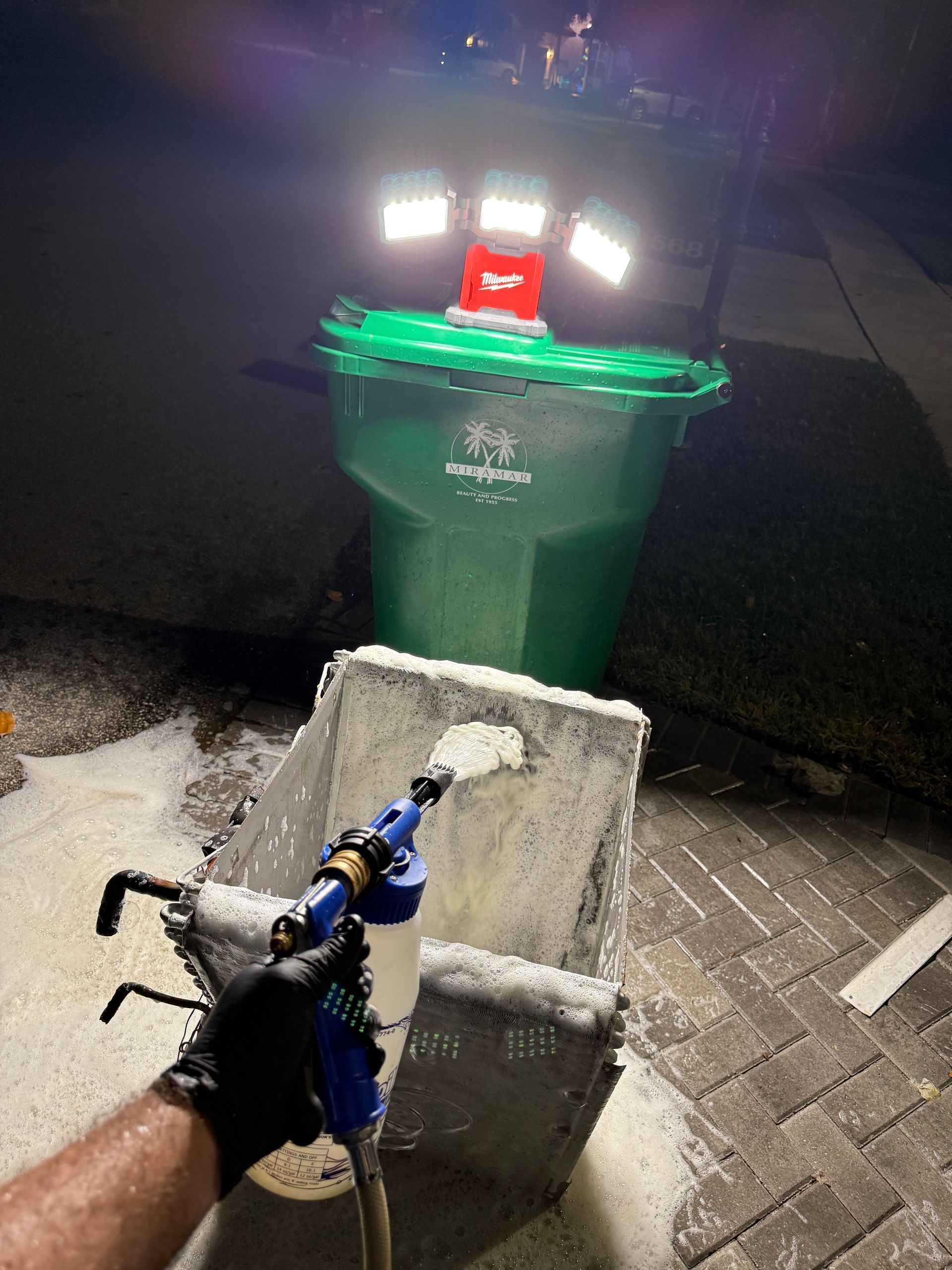 A person wearing a black glove uses a foam sprayer to clean a trash can at night, illuminated by portable work lights.