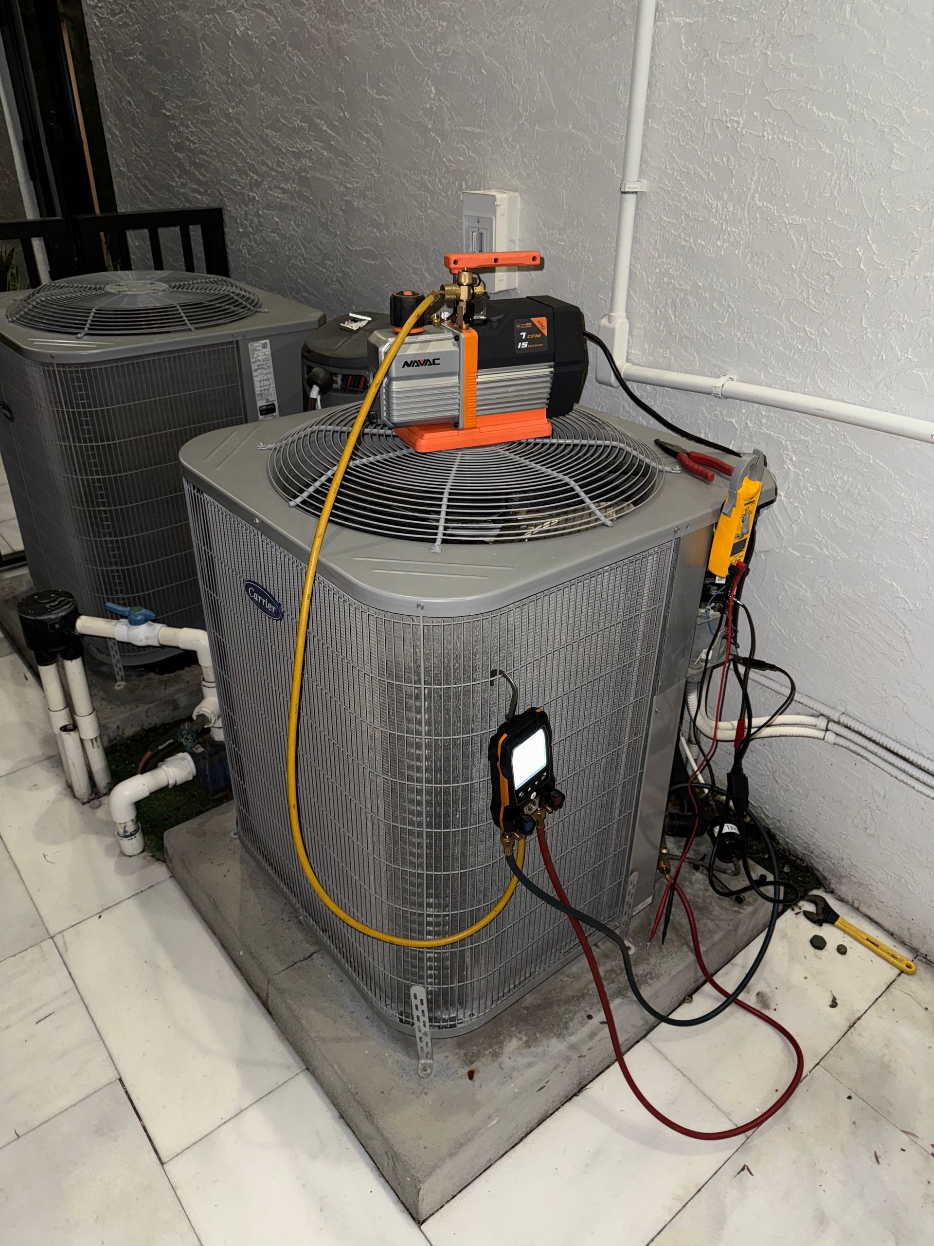 A vacuum pump sits on top of an outdoor HVAC condenser unit, connected by hoses and a digital gauge for servicing.