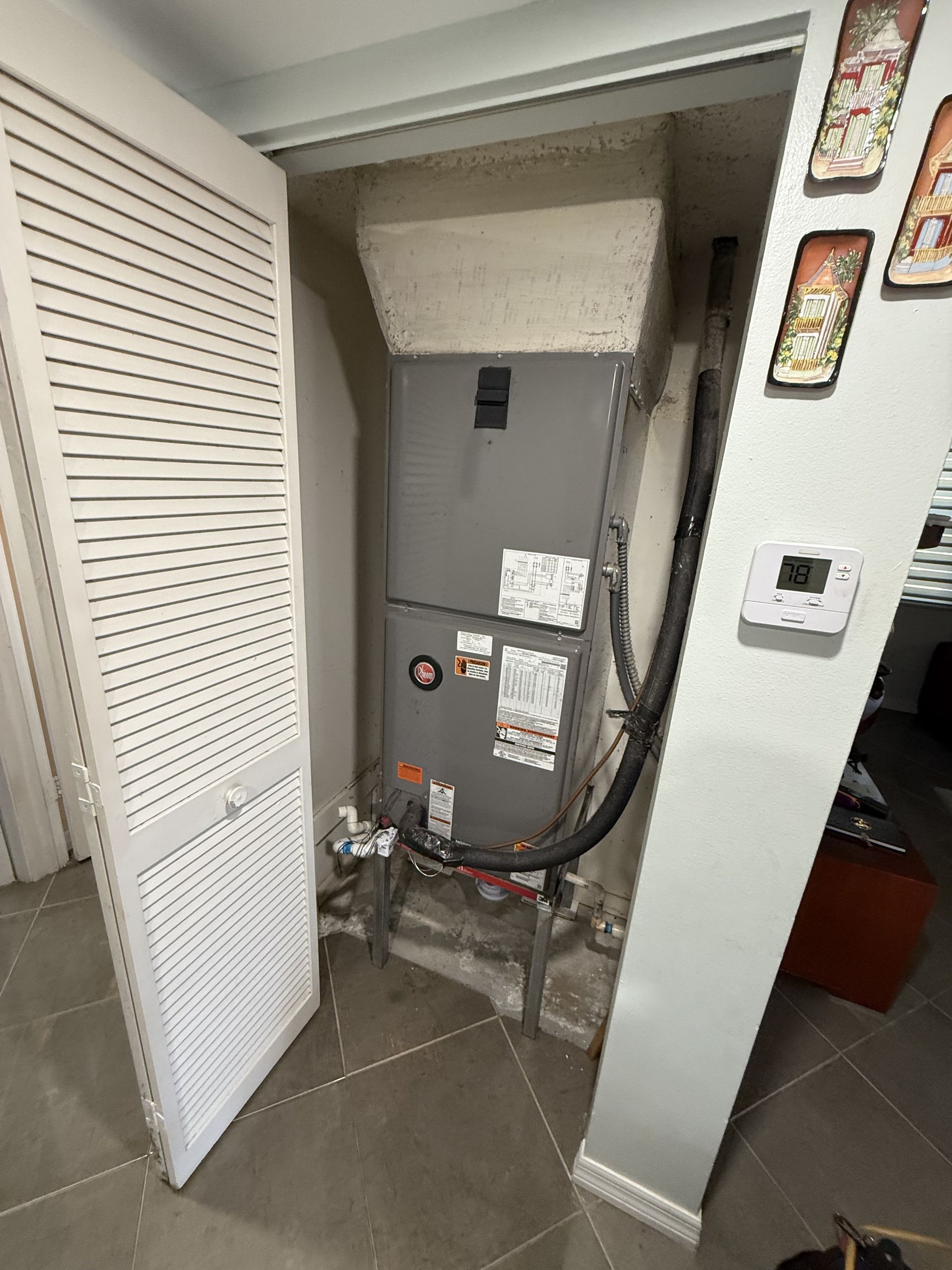 A grey HVAC furnace unit installed in a closet next to a white louvered door and a wall-mounted thermostat.