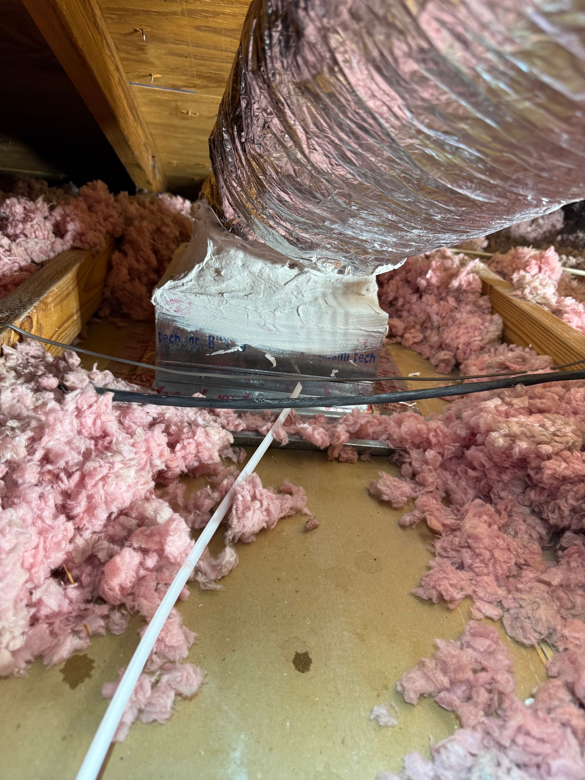 A flexible air duct connected to a metal duct box, sealed with white mastic, resting on attic insulation and wooden joists.