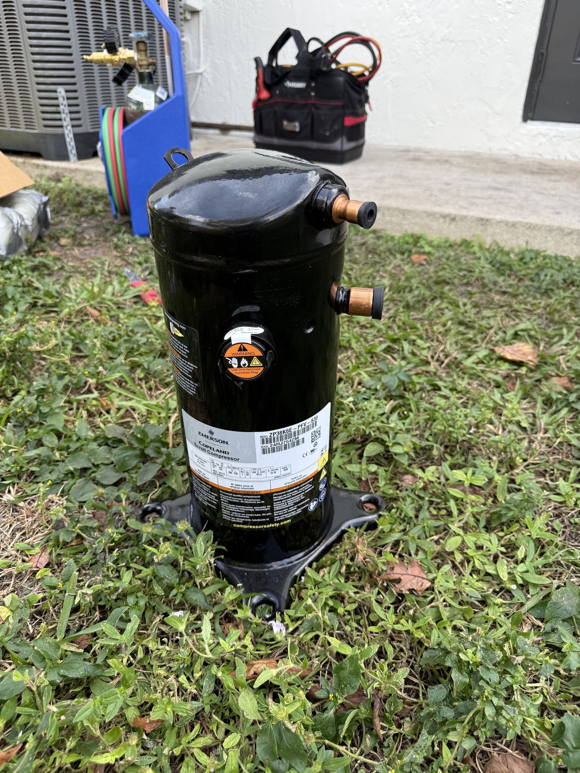 A black HVAC scroll compressor sits on a patch of grass near a toolbox and an air conditioning unit.