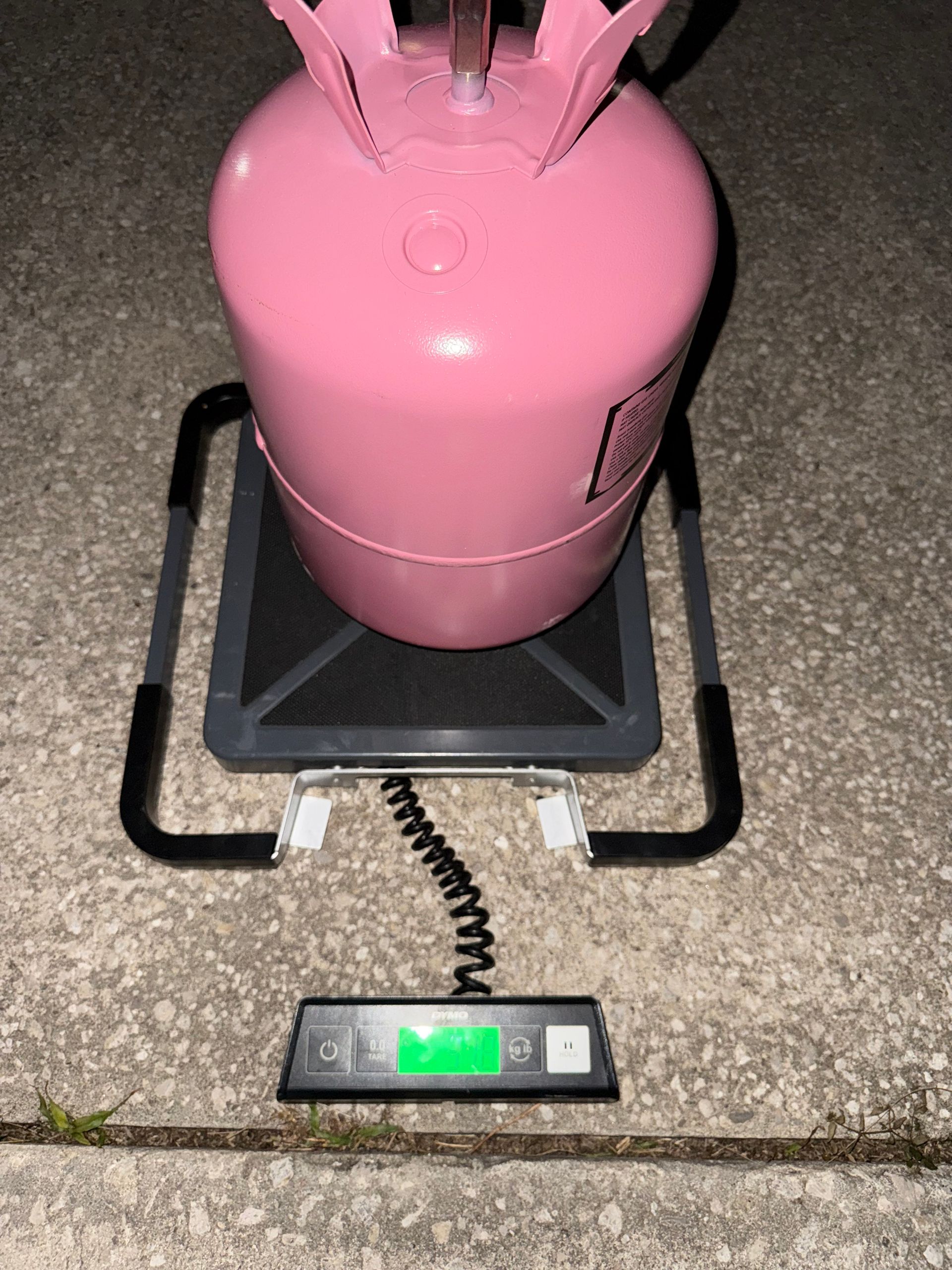 A pink refrigerant cylinder sits on a black digital scale on a concrete surface.