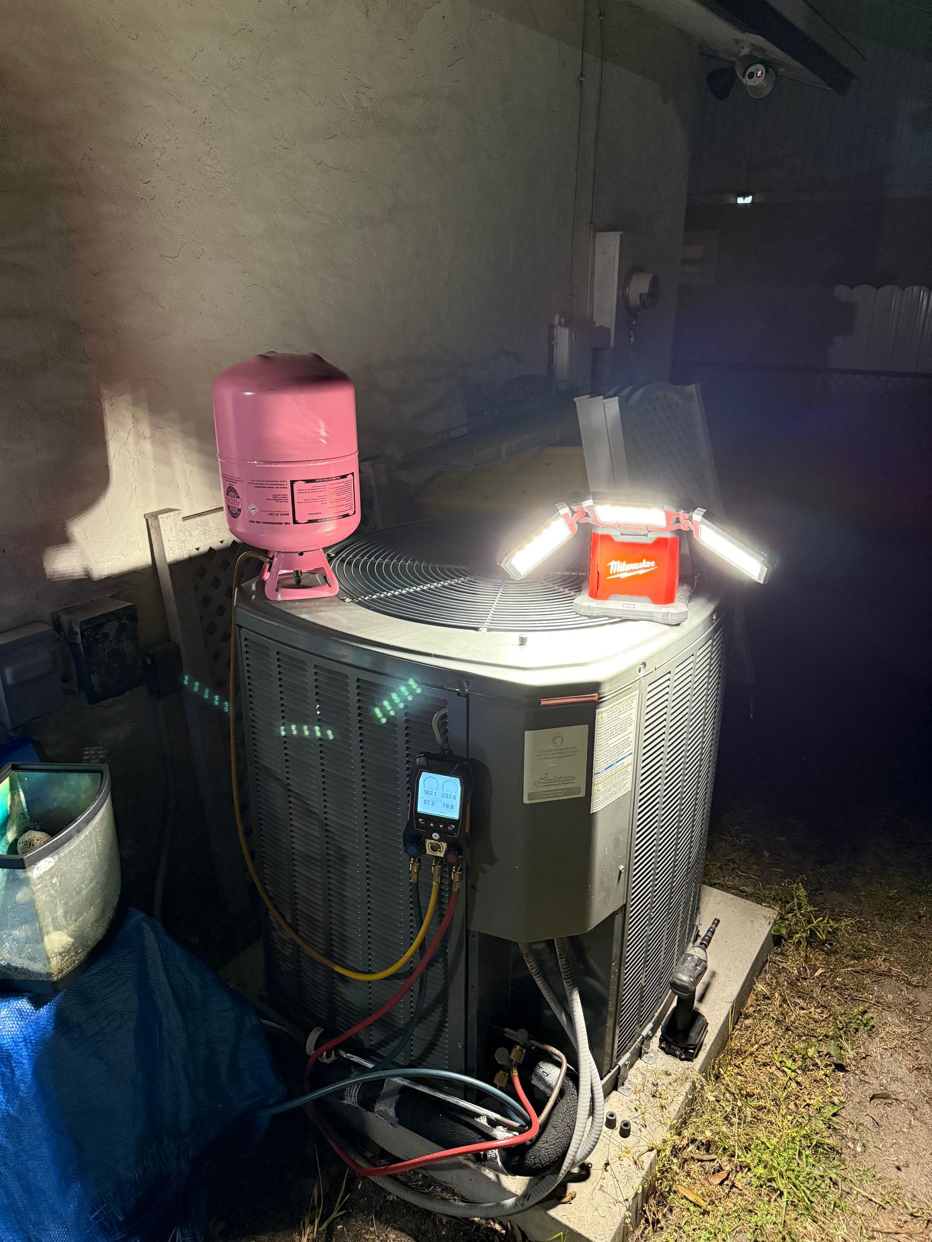 A technician’s work light illuminates an air conditioning unit at night with a refrigerant cylinder on top.