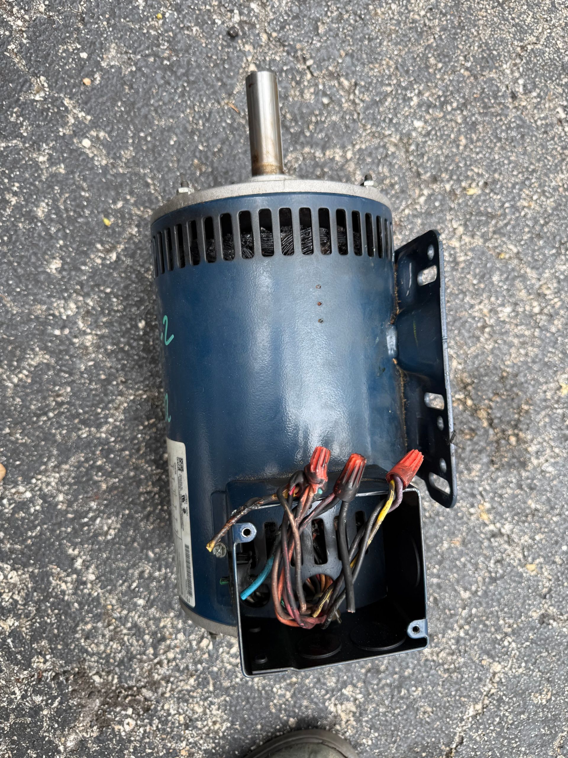 A blue electric motor with an exposed wire junction box and a metal drive shaft, resting on a gray paved surface.