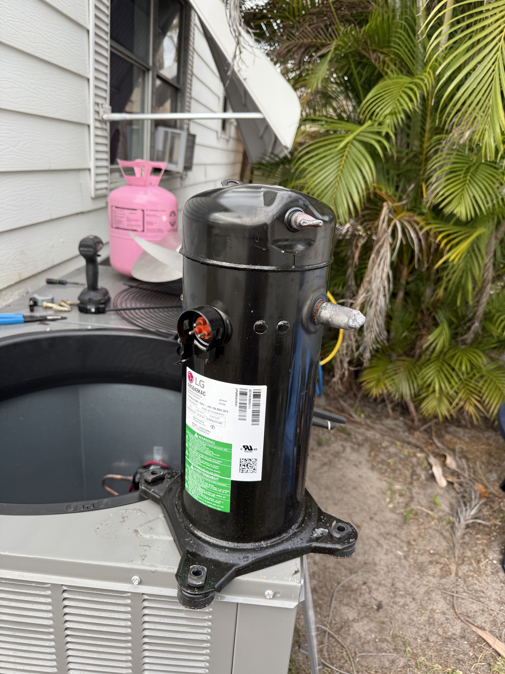 A black replacement HVAC compressor sits on top of an outdoor air conditioning unit next to a pink refrigerant tank.
