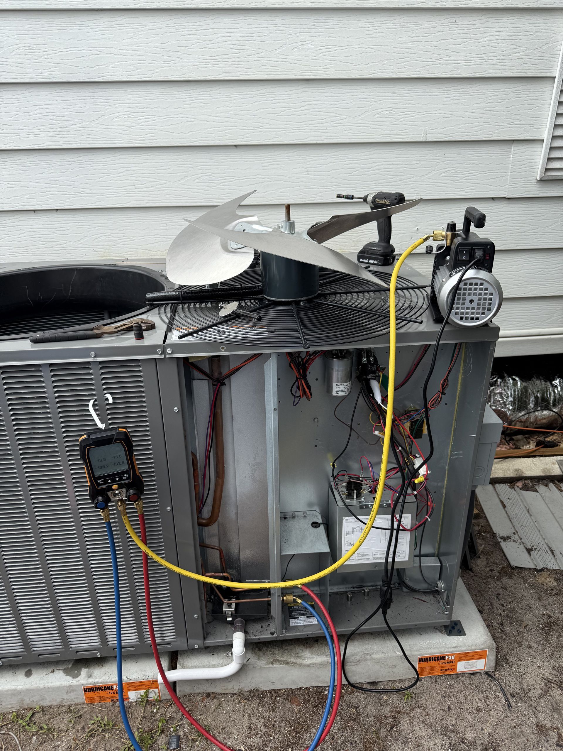 An outdoor HVAC unit under repair with a vacuum pump attached and a manifold gauge set hung on the side panel.