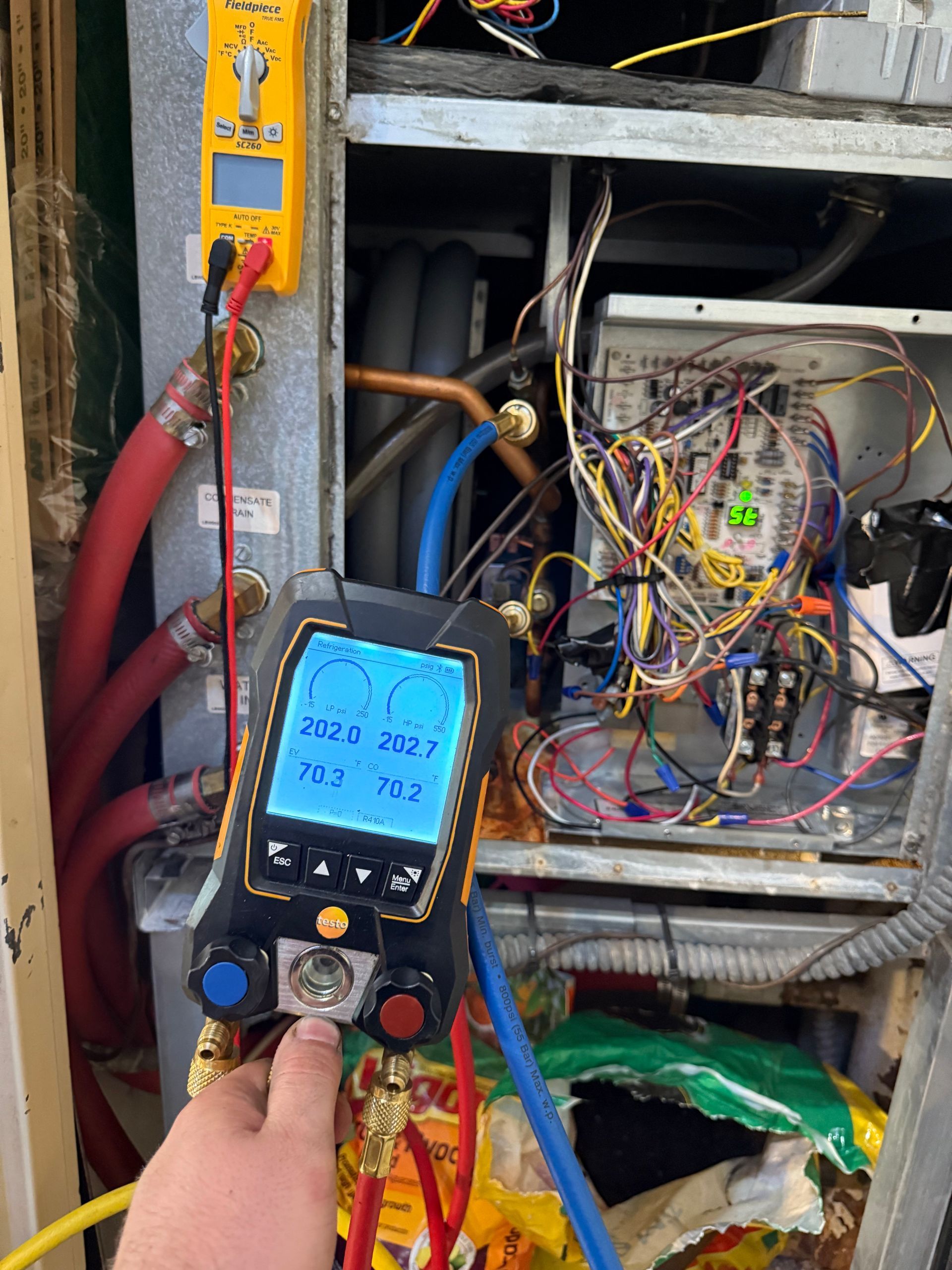 A technician’s hand holds a digital HVAC manifold gauge connected to an open air handler unit with exposed wiring.
