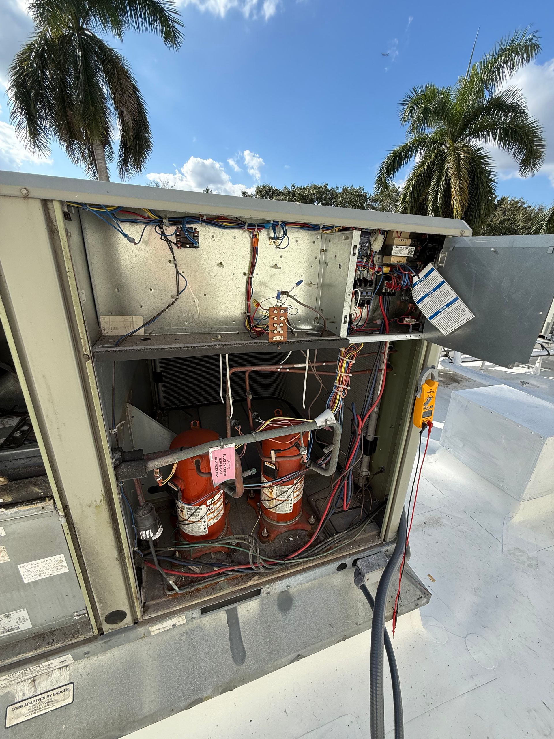 Open HVAC unit on a rooftop, showing two cylindrical compressors and exposed wiring against a blue sky with palm trees.
