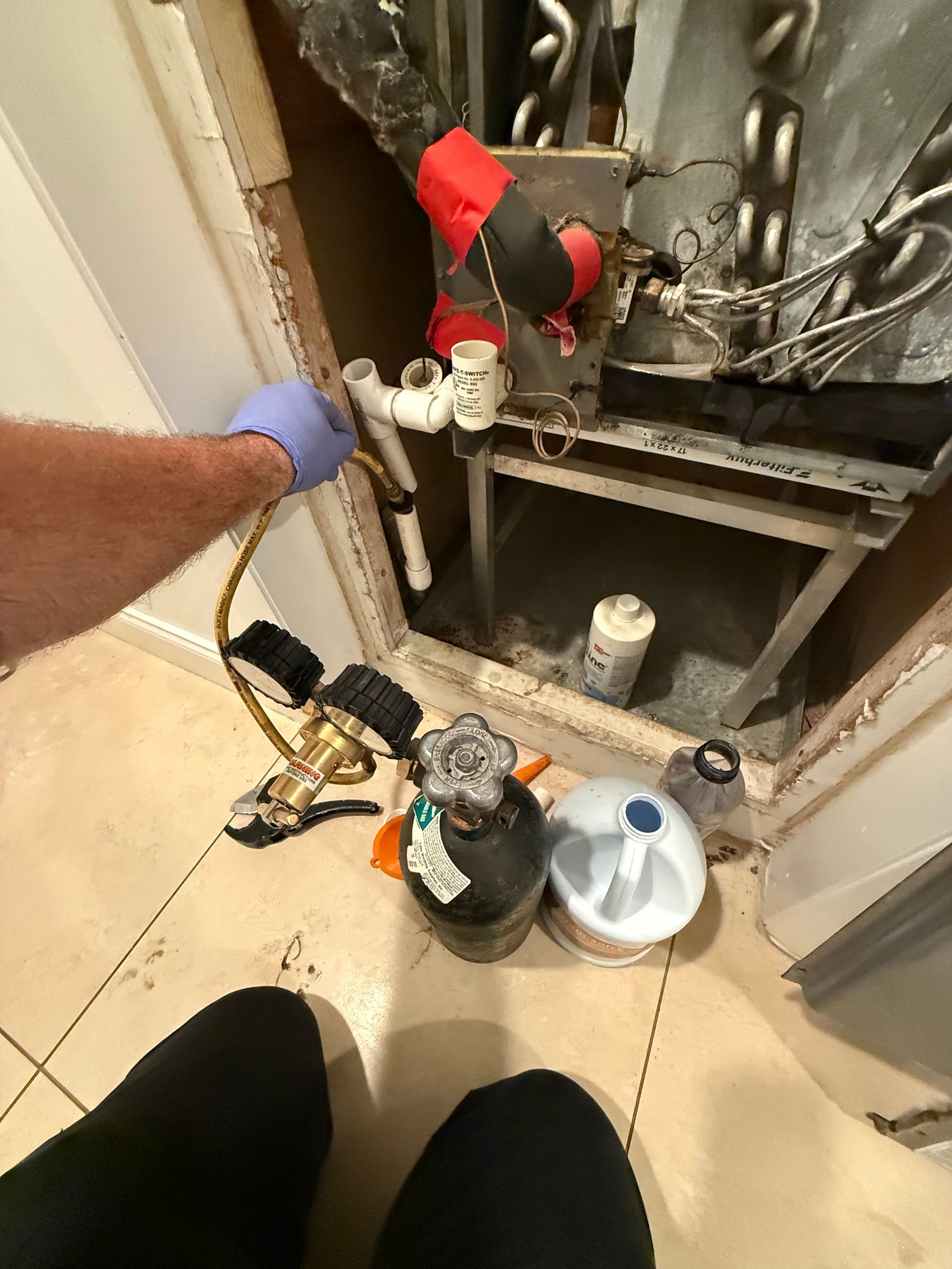 A hand in a blue glove connects a pressure gauge hose to the PVC drain pipe of an indoor HVAC unit.