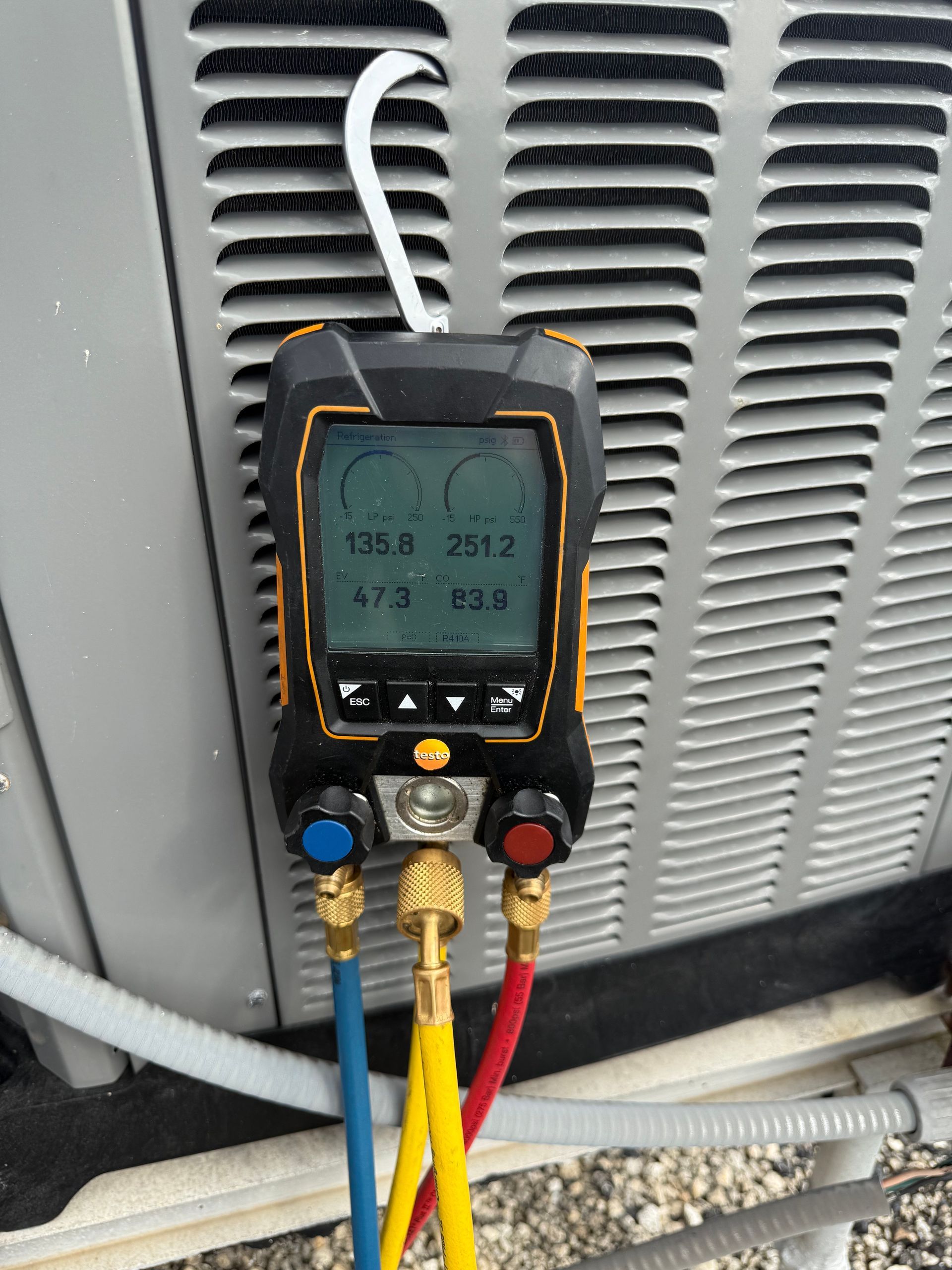 Digital manifold gauge measuring pressures and temperatures on an outdoor HVAC condenser unit.