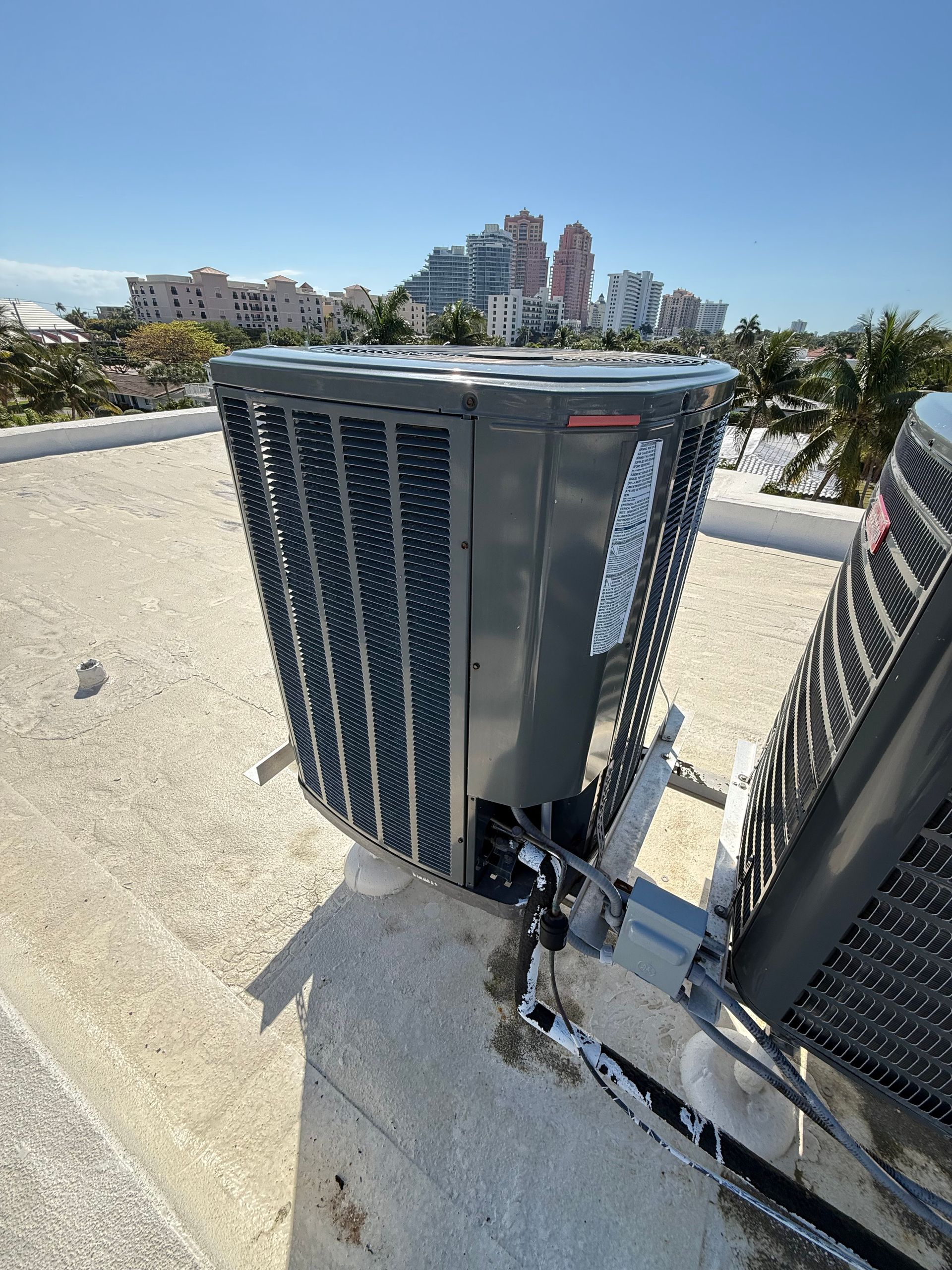 Two gray industrial air conditioning units positioned on a flat, gravel-covered roof under a bright blue sky.