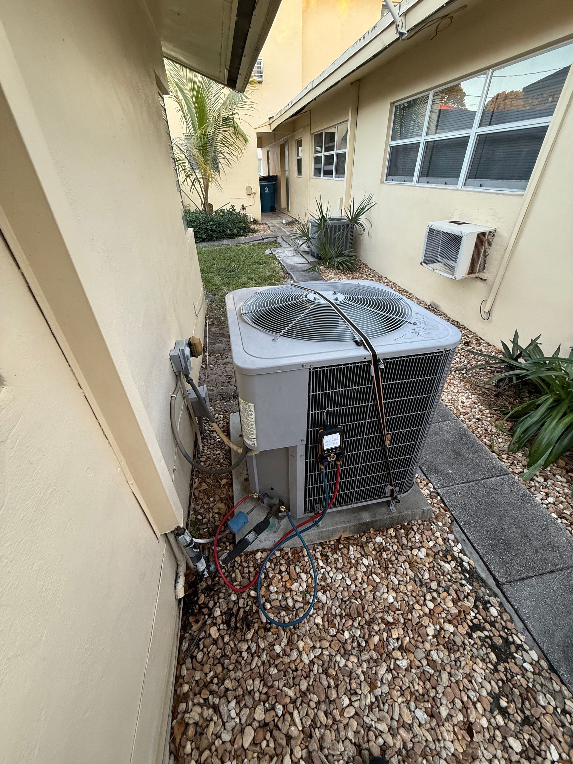 A gray HVAC condenser unit sits on gravel between two beige exterior building walls in an outdoor passageway.