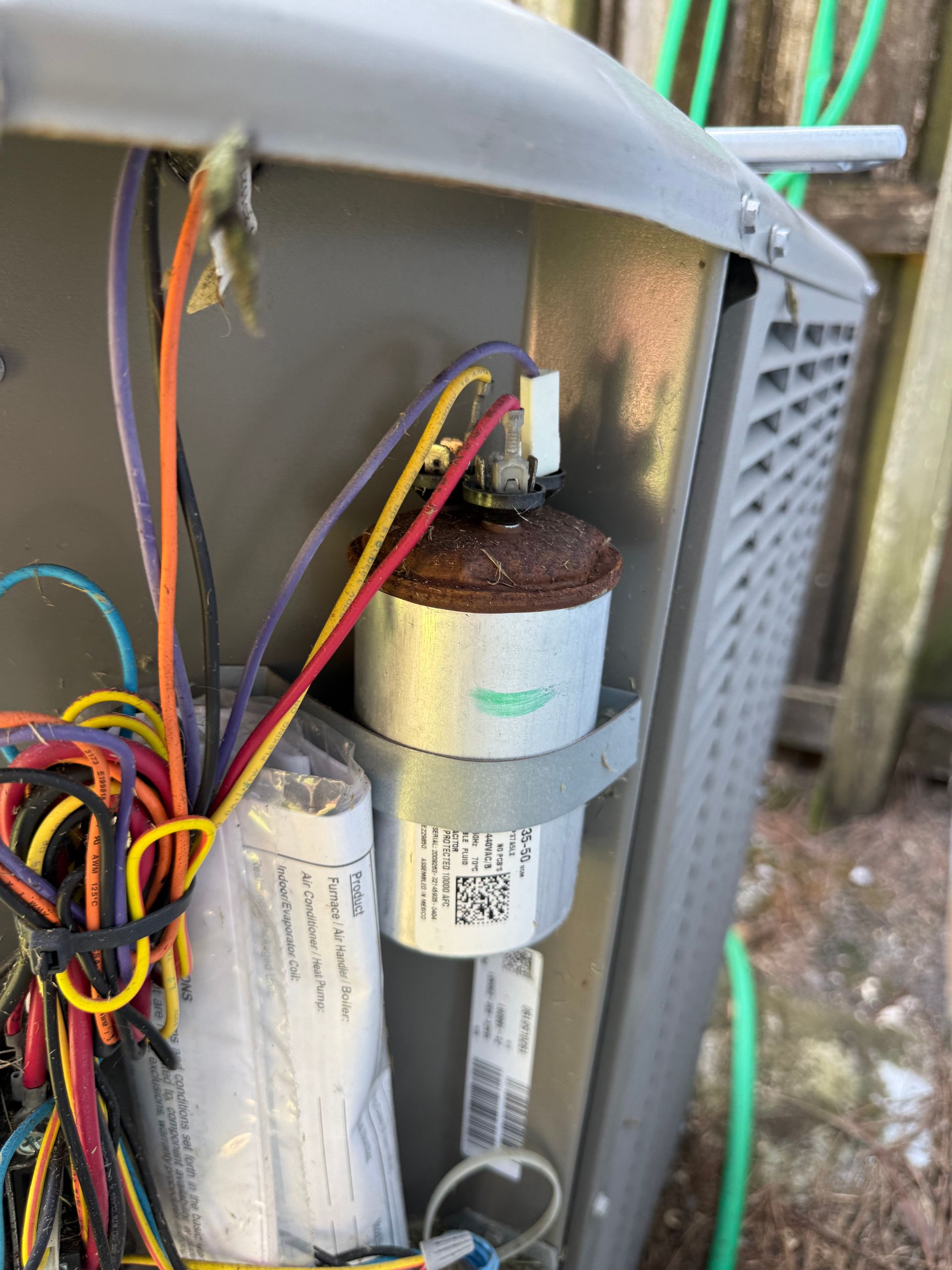 A cylindrical metal capacitor with a rusted top and attached colorful wires inside an outdoor HVAC unit cabinet.