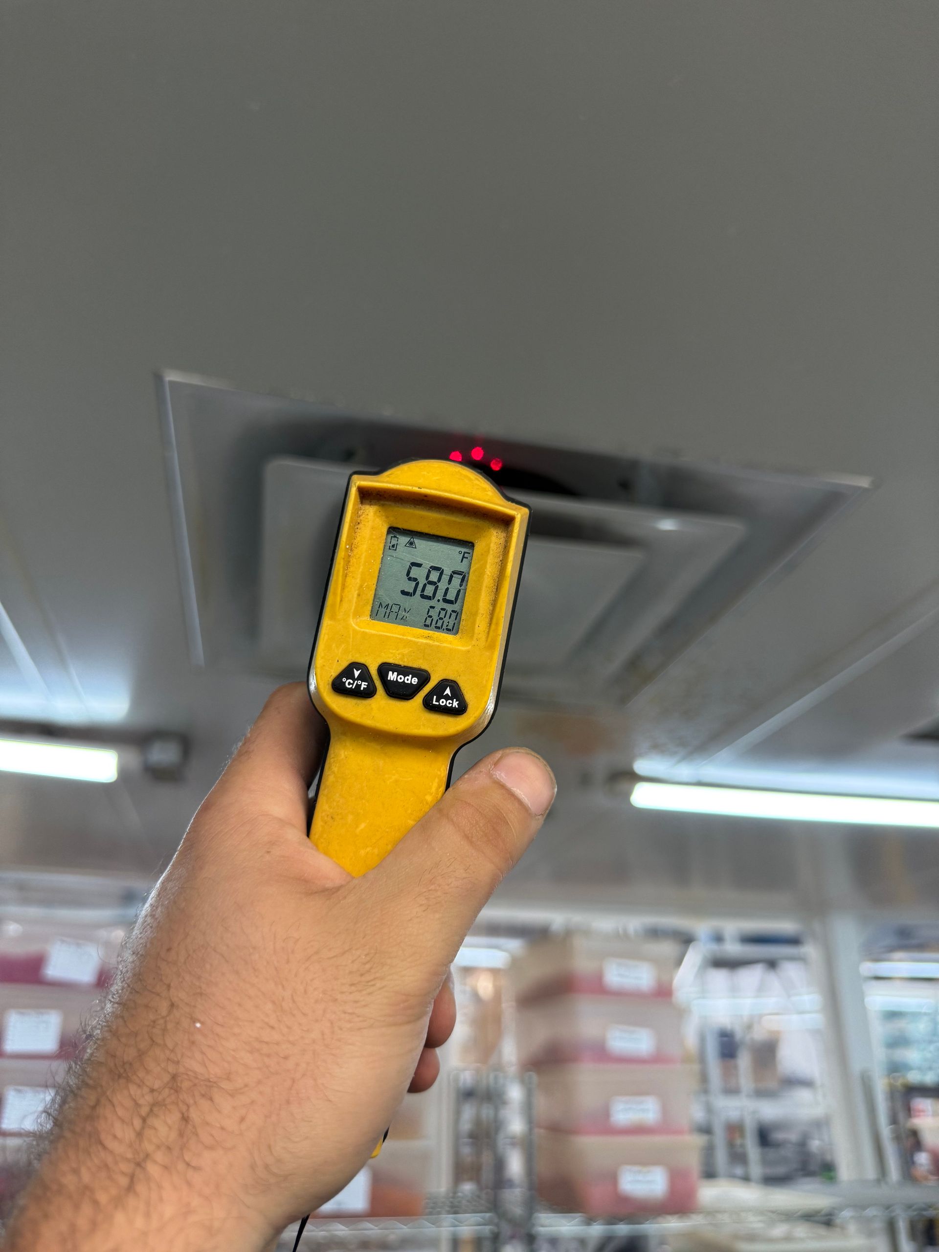 A hand holding a yellow infrared thermometer gun pointed at a ceiling air vent, displaying a reading of 58.0.