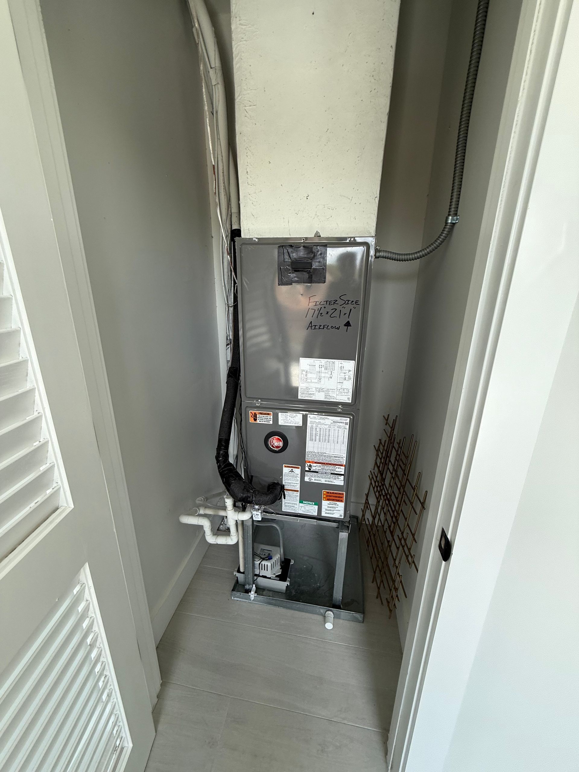 A vertical HVAC unit inside a small, white-walled utility closet with a louvered door to the left.