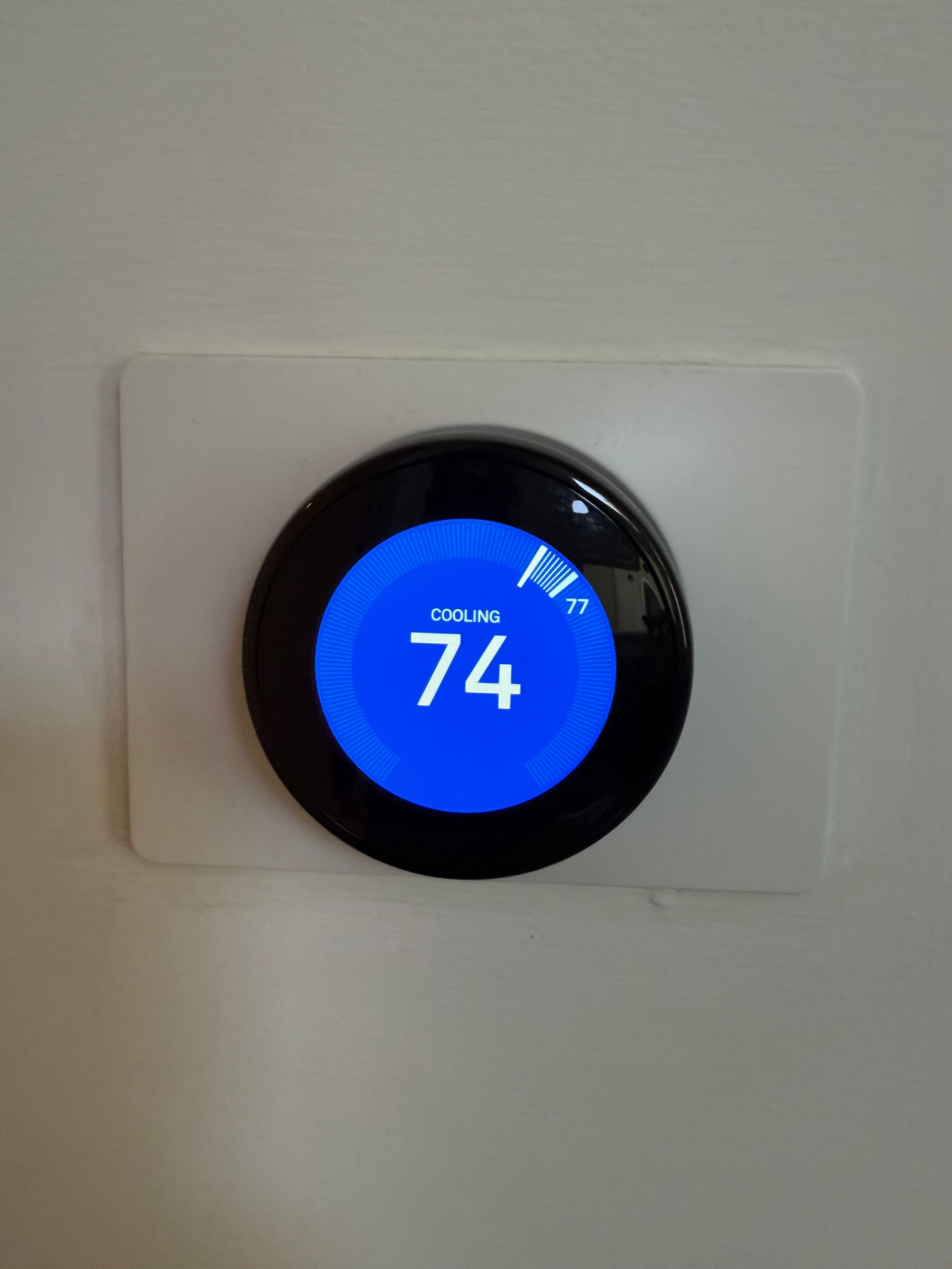 A Nest smart thermostat on a white wall displays a blue screen with the current indoor temperature of 74 degrees.