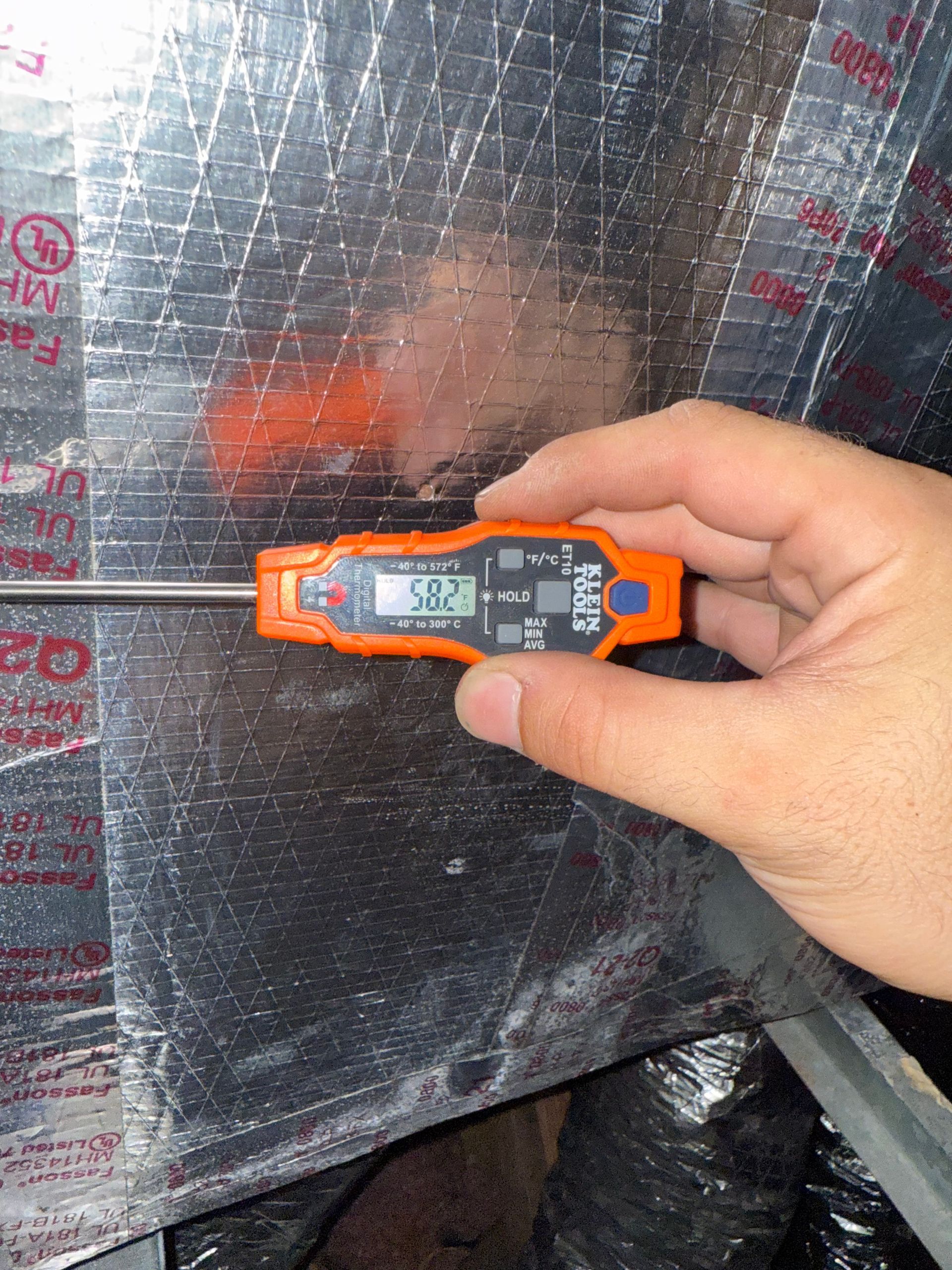 A hand holds an orange digital thermometer displaying 58.2°F, inserted into a silver-insulated HVAC air duct.