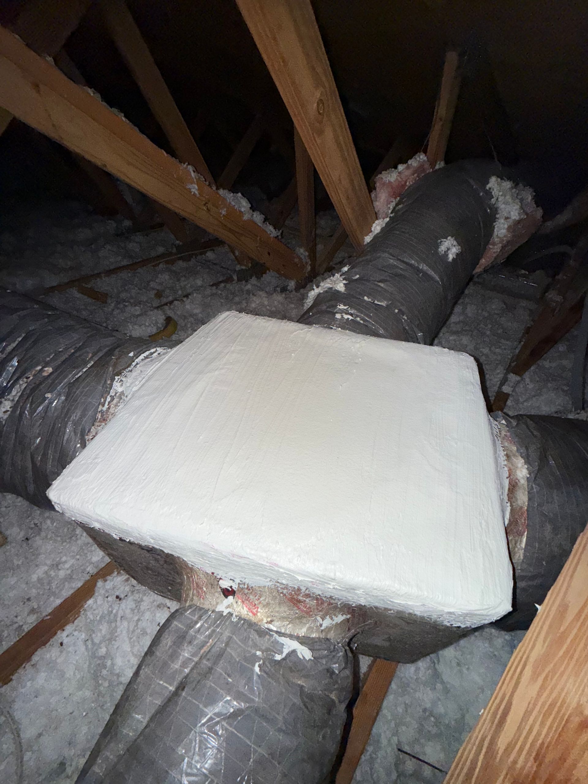 An attic view showing a duct junction box covered with a thick, white foam insulation board, surrounded by gray ducts.