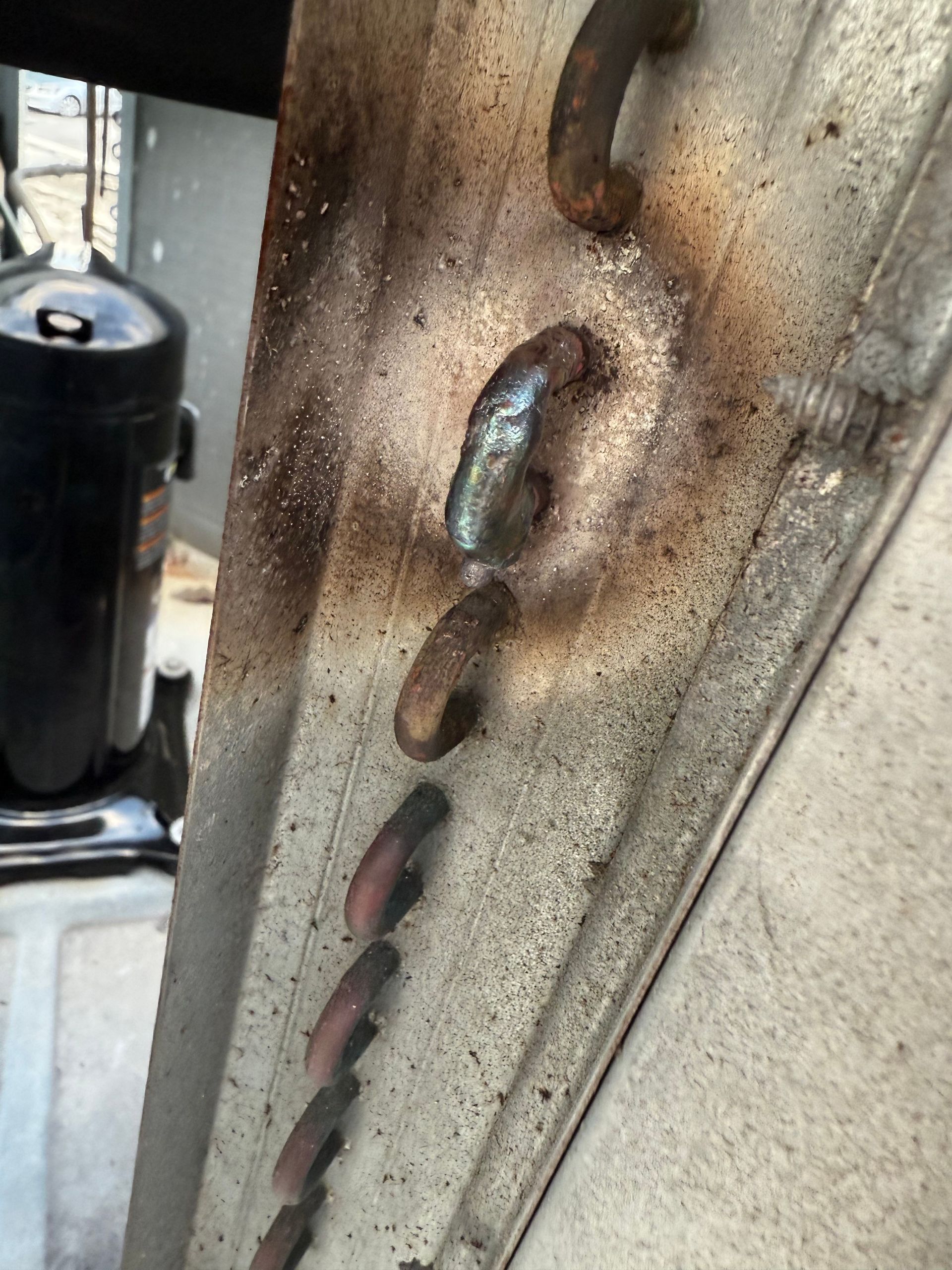 A close-up of a damaged, rusted HVAC condenser coil with a silver-soldered repair patch on one of the copper U-bends.
