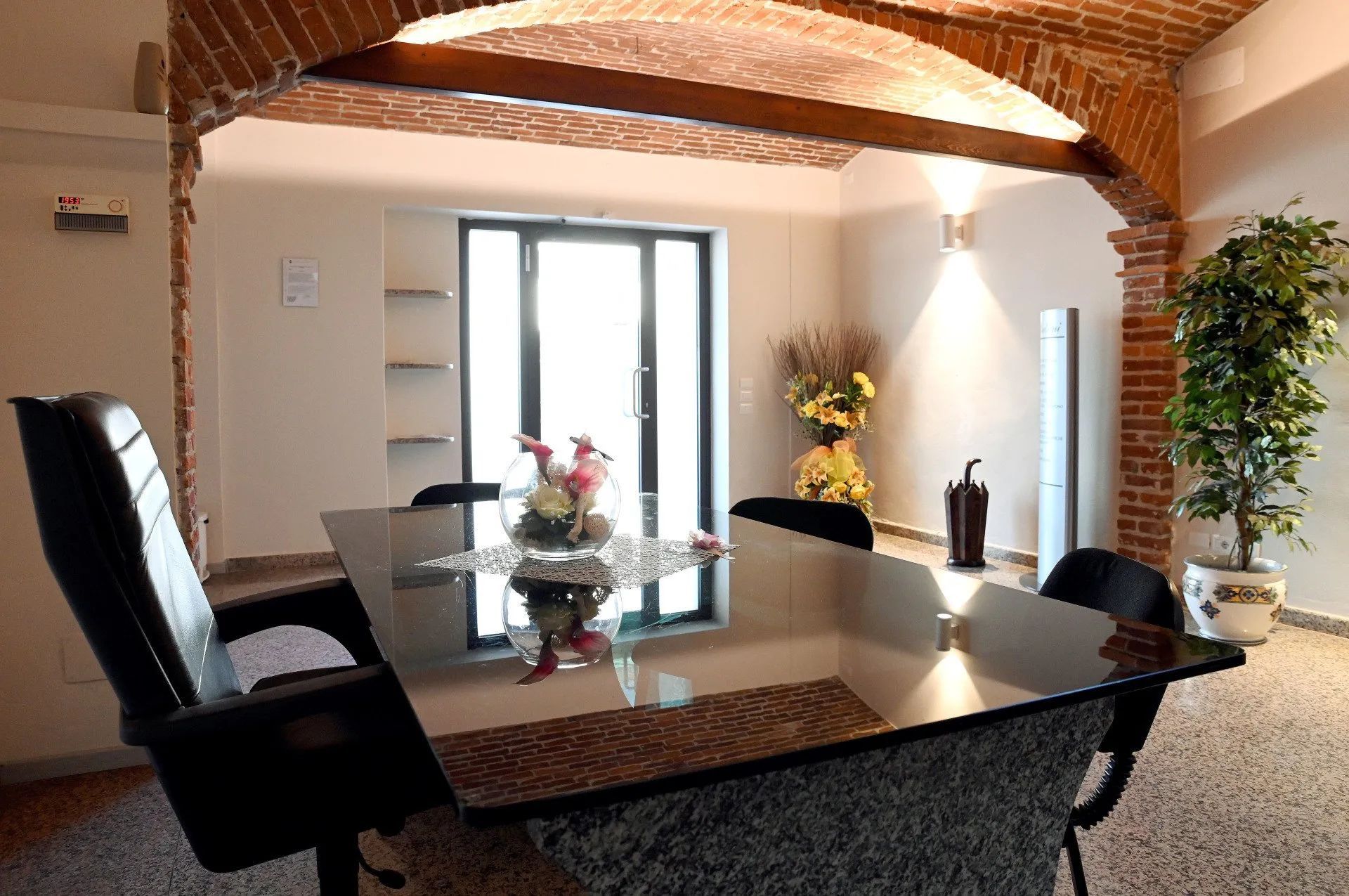 Ufficio con tavolo in vetro, sedie e arco in mattoni. Finestra e pianta in vaso visibili.