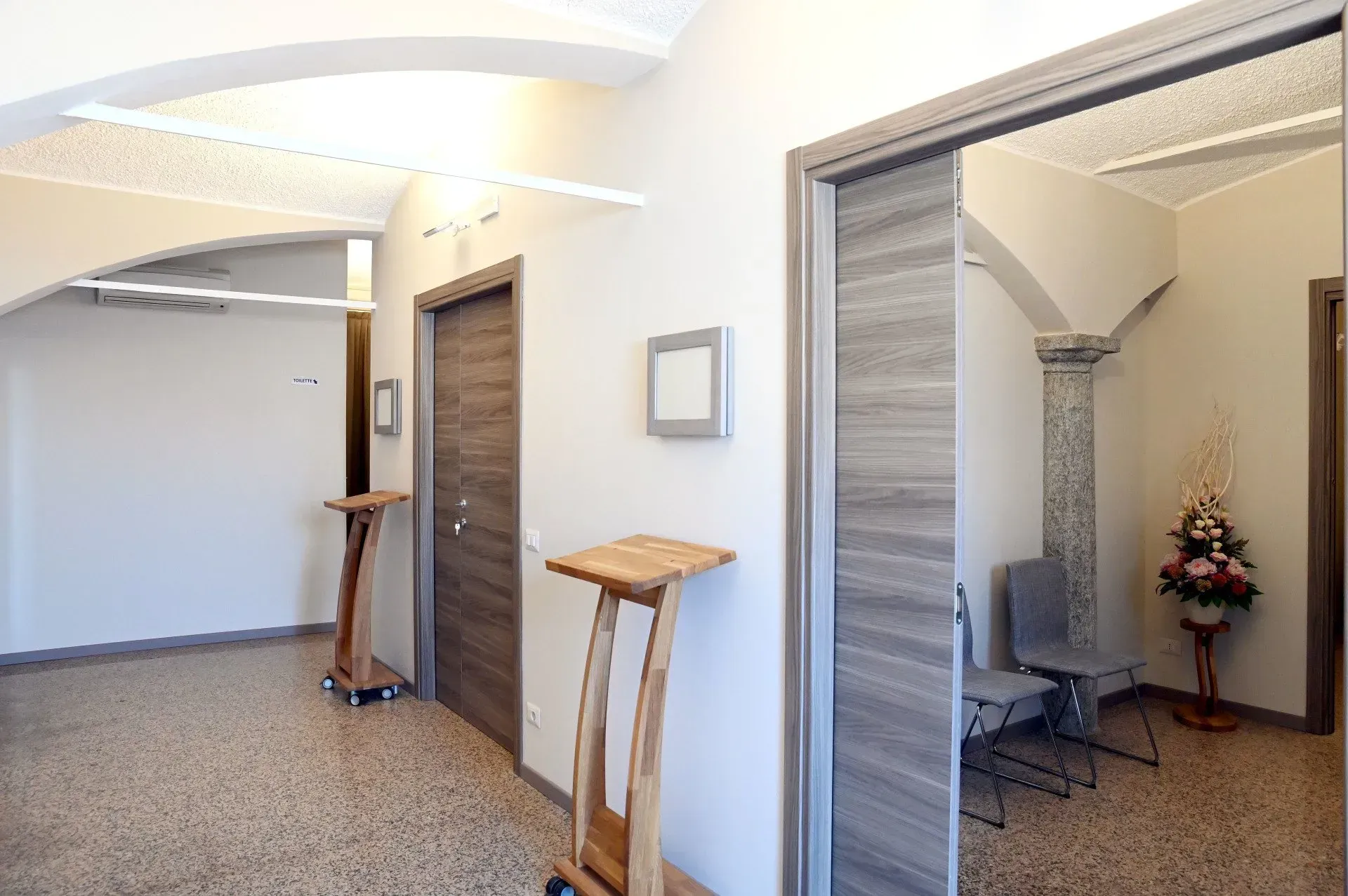 Corridoio con pavimento a macchie beige, porte in legno e portali ad arco, due podi e una composizione floreale.
