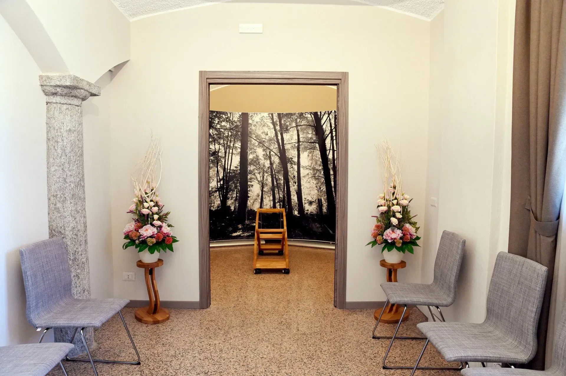 Interno con sedie, composizioni floreali e una porta aperta che rivela uno sfondo boschivo.